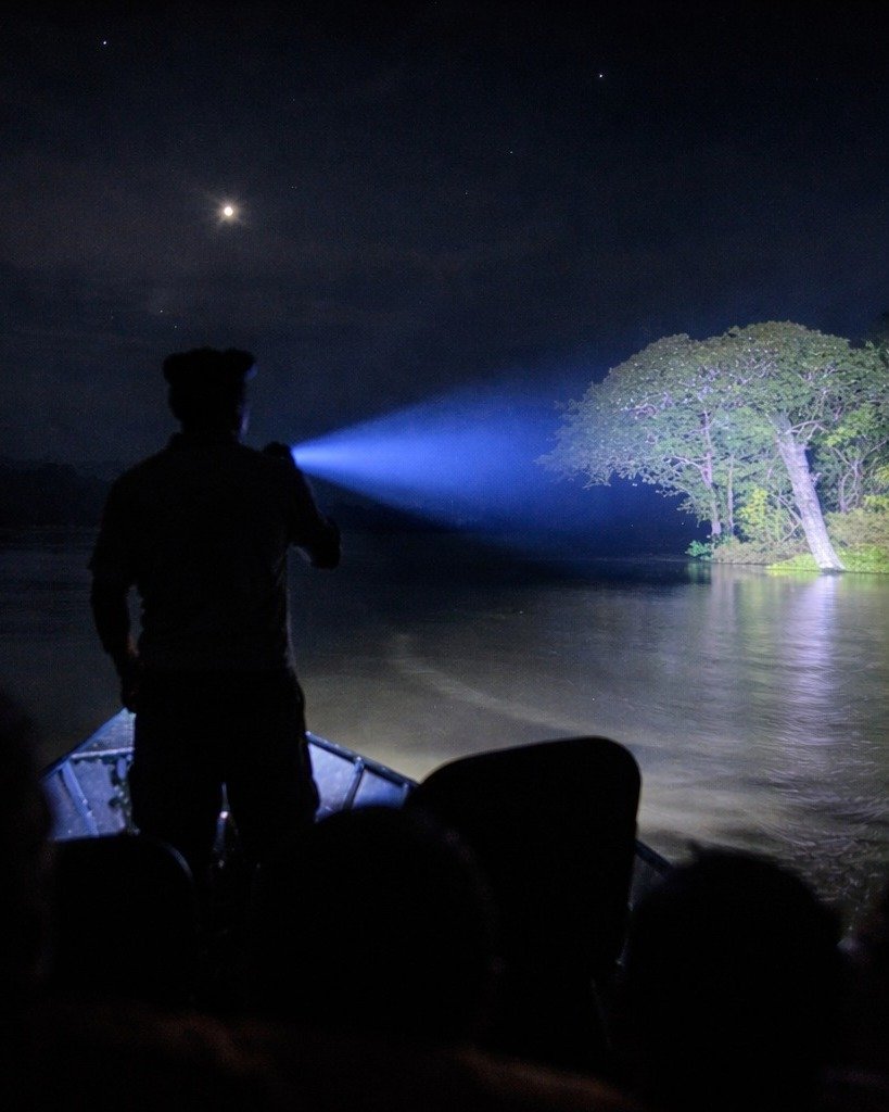 Spotlight on the river, stars overhead — nocturnal Amazônia at its most electric. A silhouetted guide standing on the bow of a small boat shines a powerful spotlight across a dark river toward a large tree at night during a Juma Amazon Jungle Lodge night tour, with a crescent moon visible above