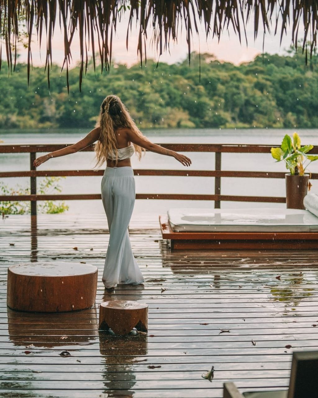 Arms wide open, rain falling soft — surrender to the Amazon. A woman in white stands arms outstretched on a rain-soaked deck at Juma Amazon Jungle Lodge, facing a calm river lined with dense green rainforest canopy under a thatched roof
