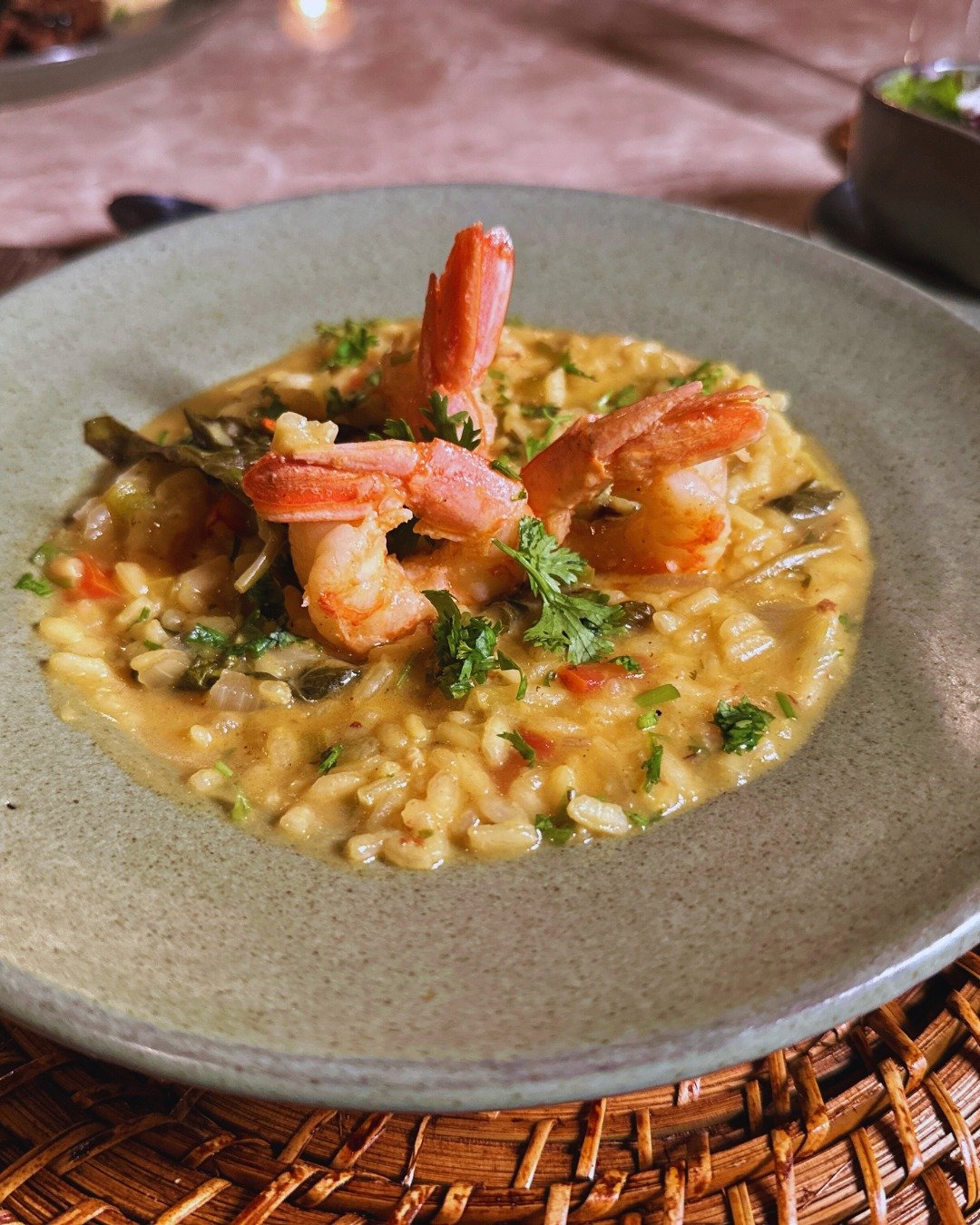 Golden, herb-flecked, and crowned with whole prawns — comfort food deep in the rainforest. A bowl of creamy shrimp risotto garnished with fresh herbs, served at Juma Amazon Jungle Lodge on a woven rattan placemat by candlelight