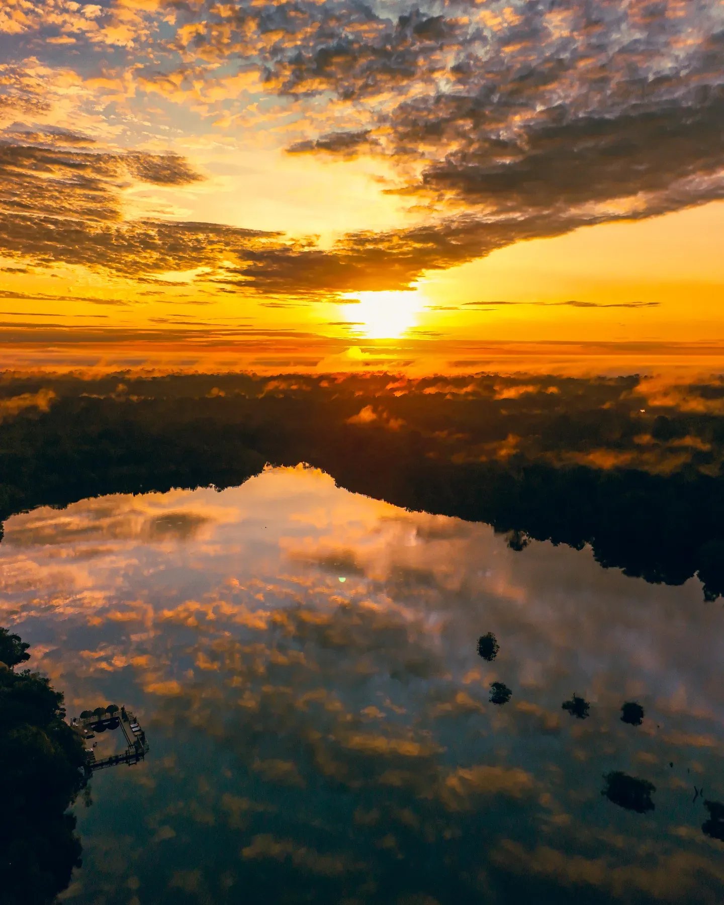 Aerial view of a blazing golden sunrise over a rainforest river at Juma Amazon Jungle Lodge, with dramatic orange clouds mirrored perfectly on the glassy water surface