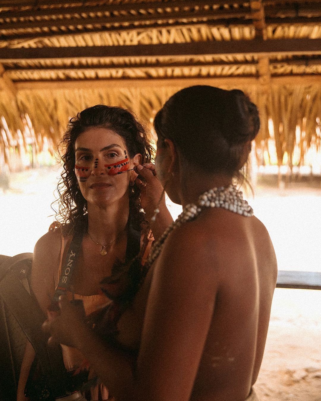 Red pigment, steady hand — a traveller receives traditional face paint from a local community member. An indigenous woman applies traditional red and black face paint to a smiling traveller inside a thatched-roof open-air hut at Juma Amazon Jungle Lodge