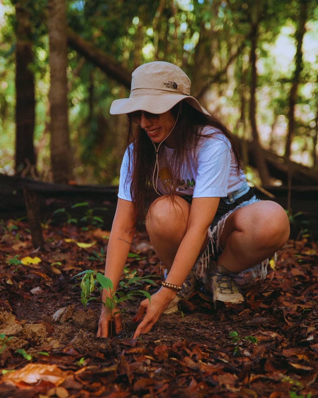Hands in the earth, giving back to the forest. A smiling woman in a wide-brim hat crouches to plant a young seedling in the forest floor at Juma Amazon Jungle Lodge, hands pressing dark soil around its roots