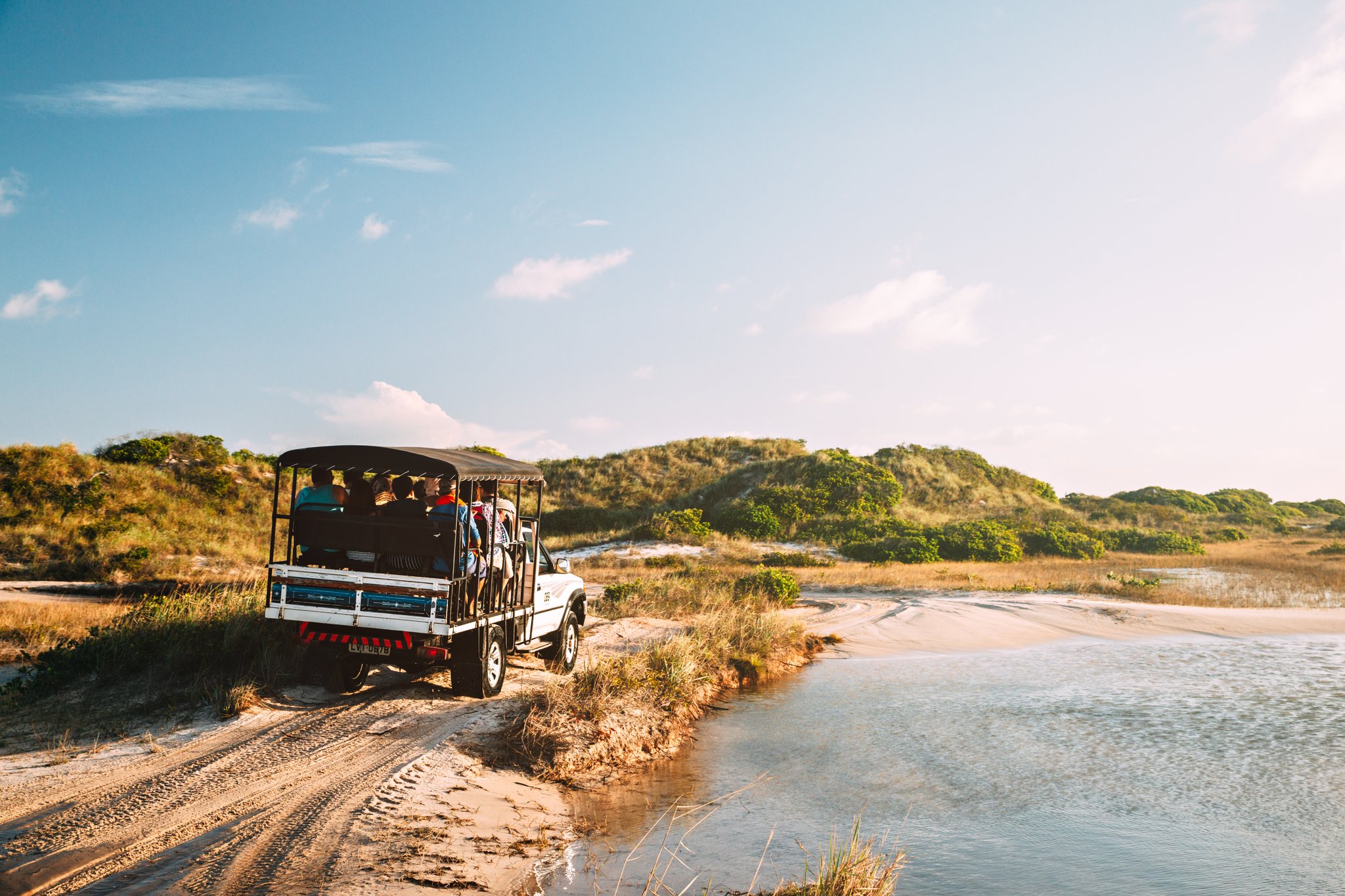 Sandy tracks, shallow water, big skies — the ride to the dunes begins. Photo: cokada An open-sided 4x4 safari vehicle carrying a group of travelers along a sandy coastal track beside a shallow tidal waterway, with green-topped dunes stretching under a blue sky — part of a Lençóis Maranhenses full itinerary transfer
