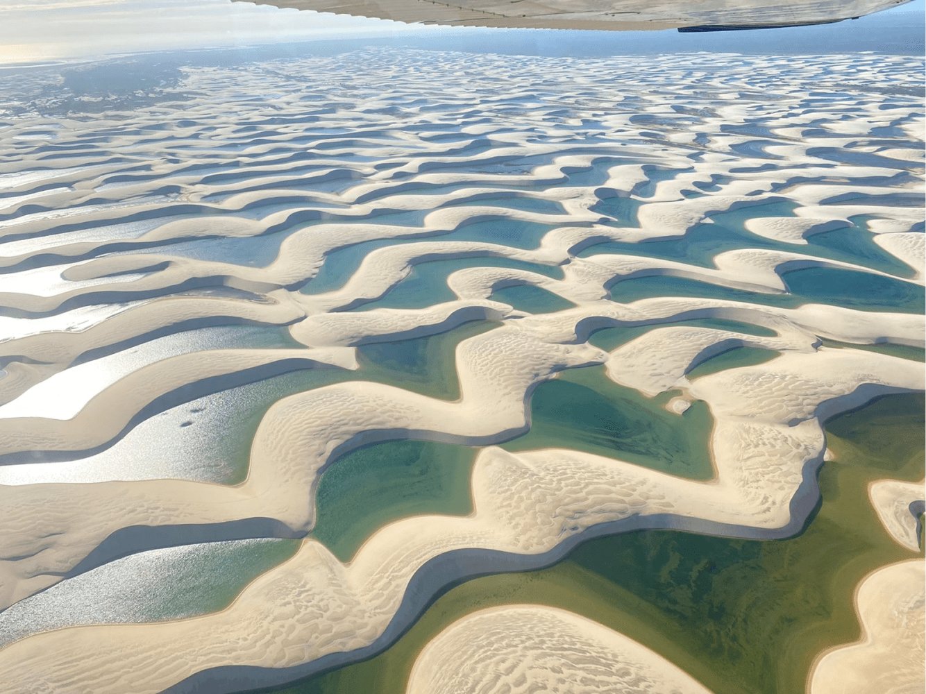 An endless mosaic of white dunes and turquoise pools, seen from the sky. Aerial view from a small aircraft over the Lençóis Maranhenses full itinerary landscape, showing sweeping white sand dunes interlaced with turquoise and emerald seasonal lagoons stretching to the horizon
