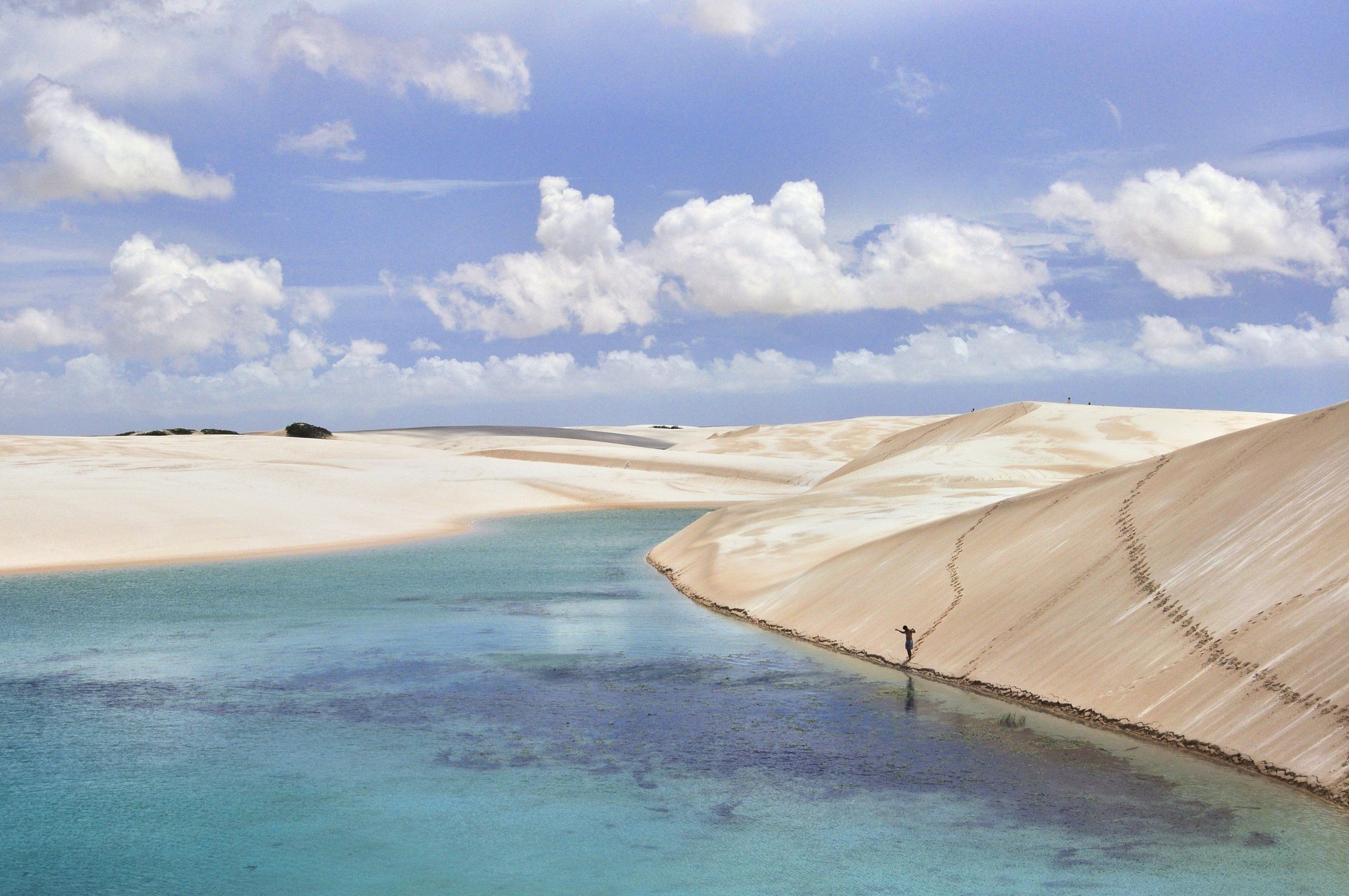 Vast white dunes, one tiny figure — scale is everything here. A lone traveller walks along the base of a towering white sand dune beside a broad turquoise lagoon in Lençóis Maranhenses Full Itinerary highlights, under a sky of dramatic clouds
