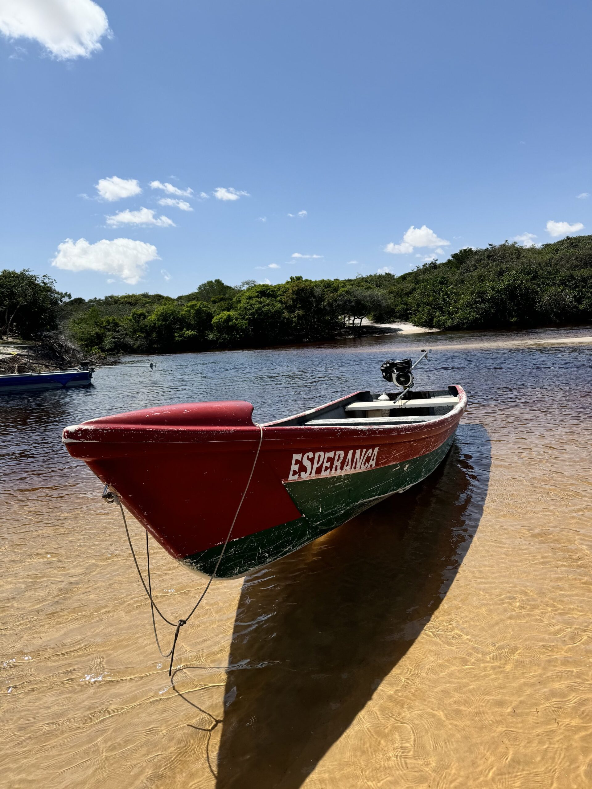 Tied up and waiting — the Preguiças River runs its own schedule. A red-and-green wooden motorboat named Esperança beached on a sandy riverbank, part of the Lençóis Maranhenses full itinerary river transfers, with dense green vegetation lining the far shore under a blue sky