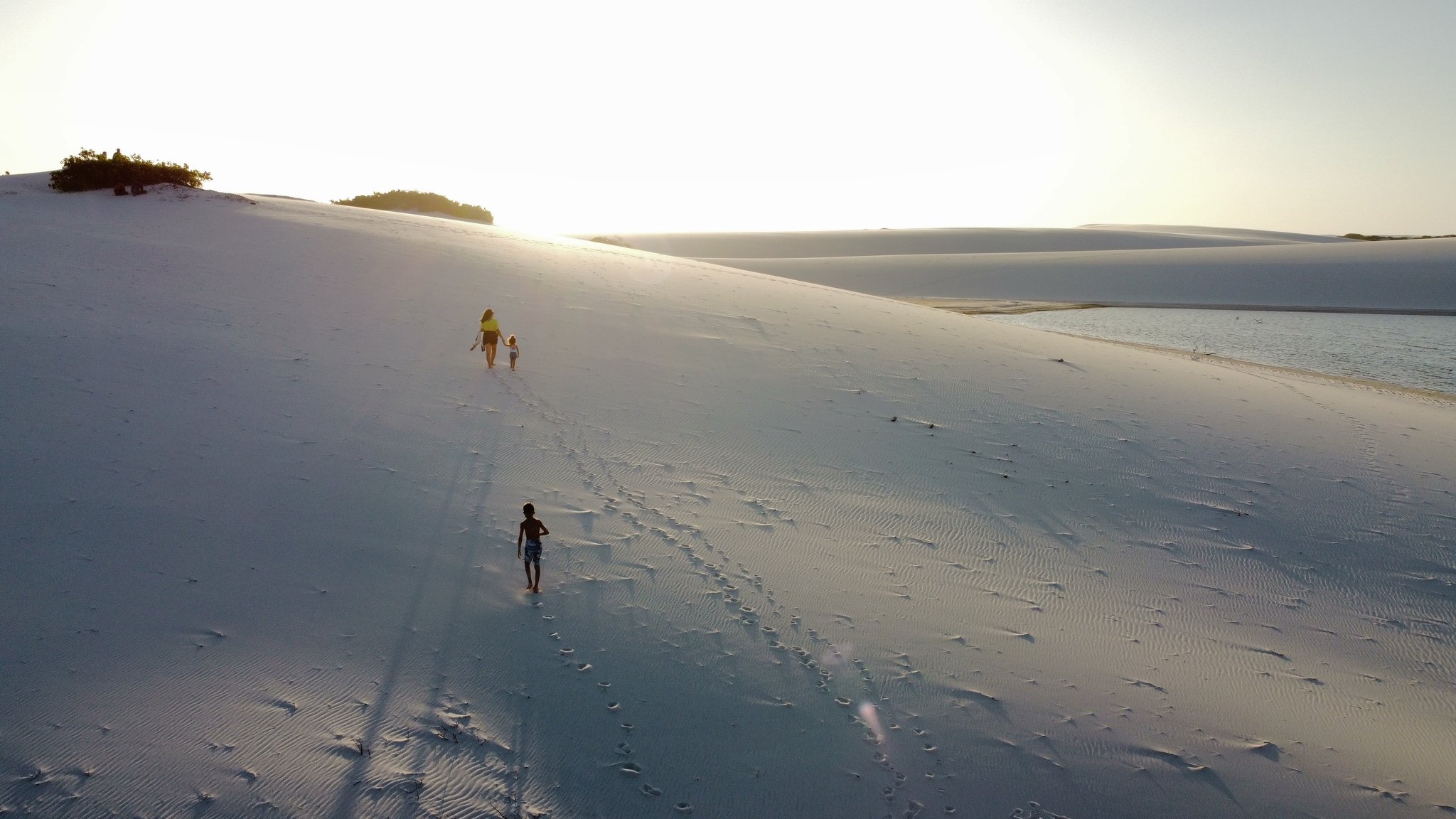 Three sets of footprints, one sweeping dune, endless horizon. An adult and two children climb the vast white sand dunes of Lençóis Maranhenses Full Itinerary territory, with a shallow lagoon glinting in golden sunset light to the right
