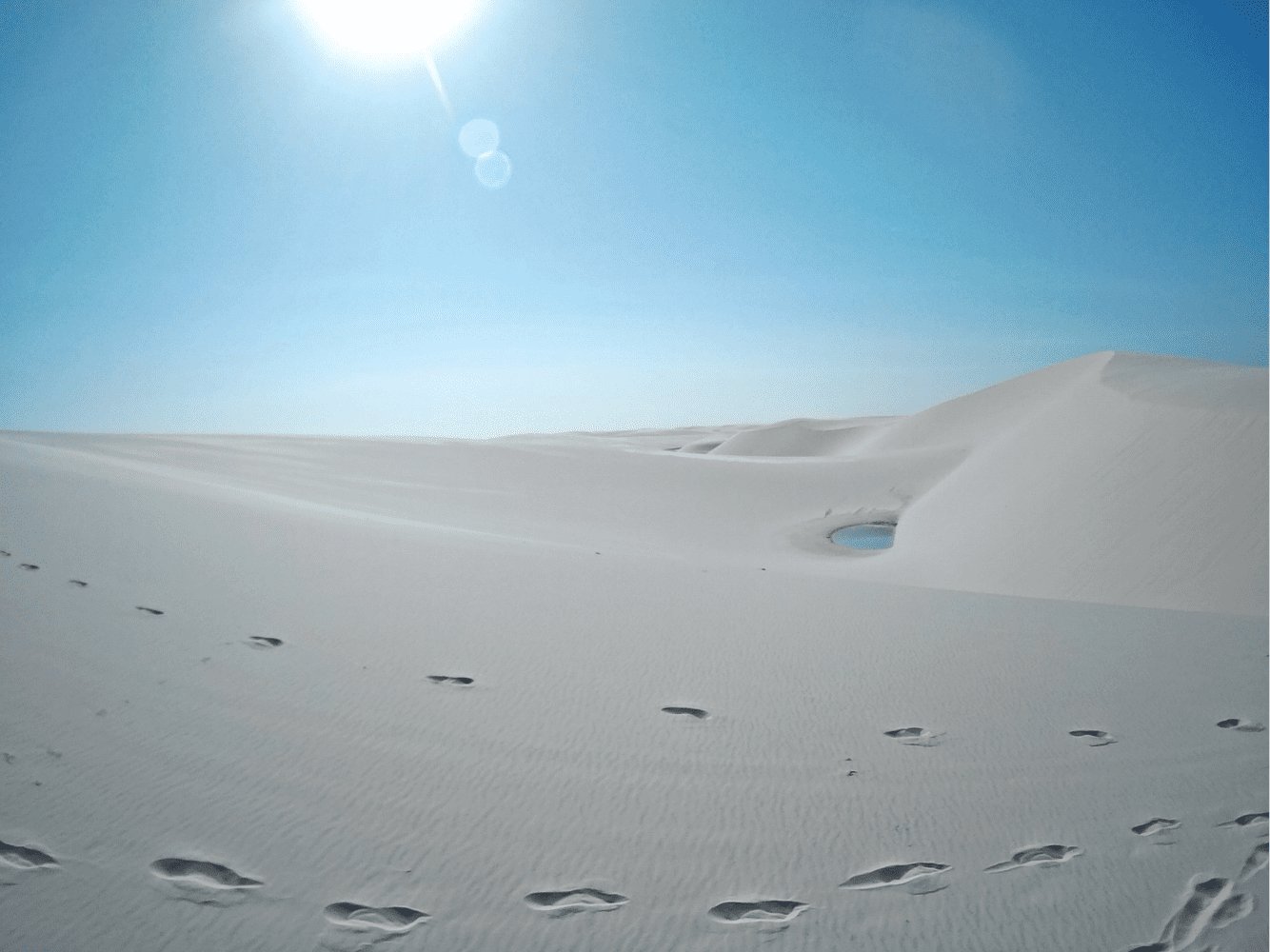 Follow the footprints — a tiny blue lagoon waits on the other side. Barefoot footprints trail across vast white sand dunes toward a small blue lagoon under a blazing midday sun in Lençóis Maranhenses full itinerary country