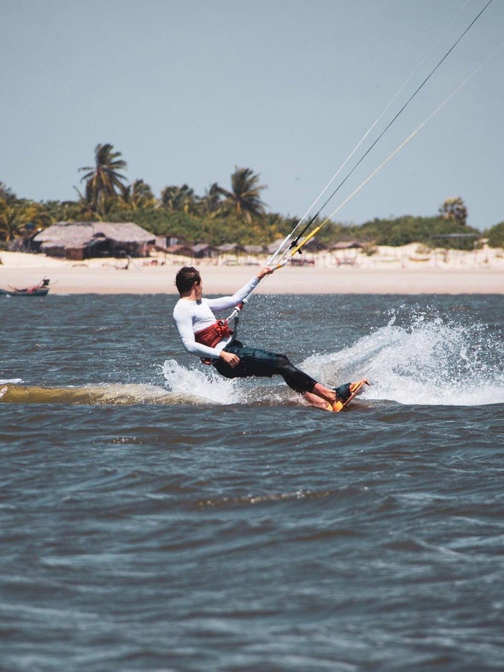 Kite lines taut, board carving spray — Atins delivers on its wind-swept reputation. A kitesurfer glides across choppy water near a white-sand beach lined with palm trees and thatched huts, part of the Lençóis Maranhenses full itinerary experience in Atins