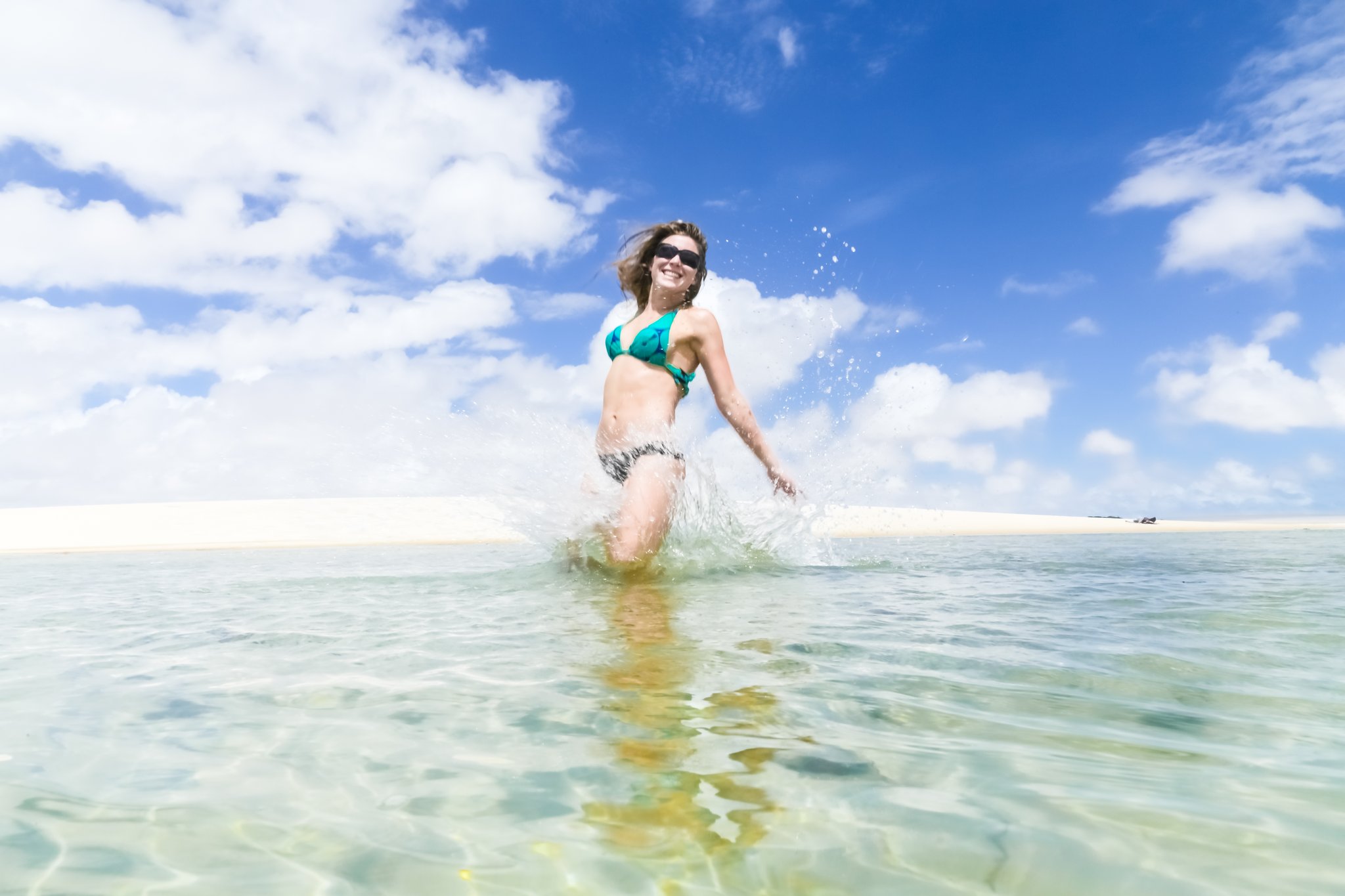 Pure joy, knee-deep in the clearest water you've ever seen. Photo: MaRabelo A smiling woman in a teal bikini splashing through crystal-clear shallow lagoon water on the Lençóis Maranhenses full itinerary, white sand dunes stretching across the horizon behind her