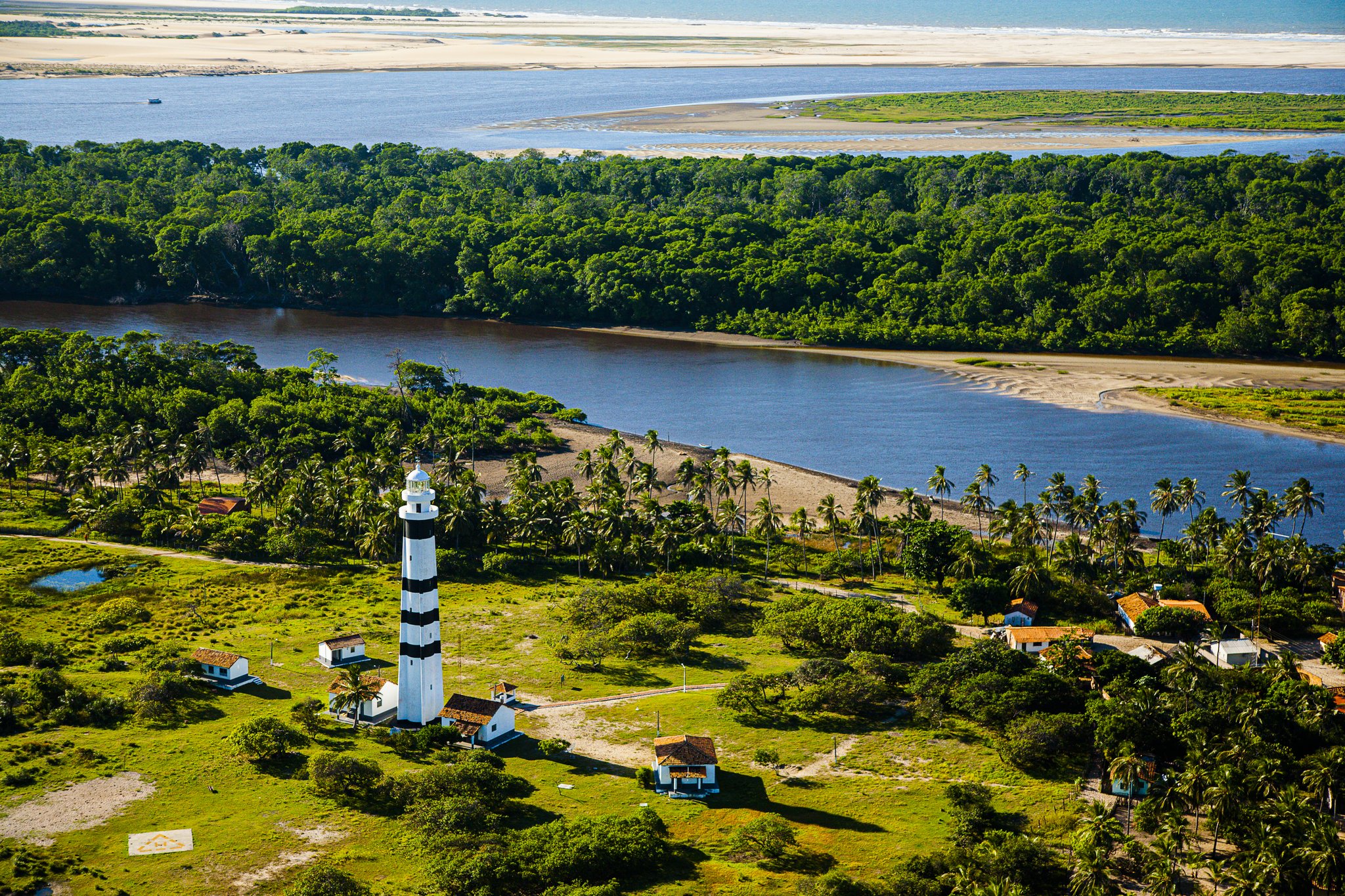 Striped sentinel at the river's edge — the Mandacaru lighthouse commands a sweeping view of the Preguiças River meeting the sea. Photo: Gabriel Boieras Aerial view of a black-and-white striped lighthouse rising above palm trees and green vegetation in the Lençóis Maranhenses full itinerary route, with the broad Preguiças River estuary, dense forest, and pale sand dunes visible in the background