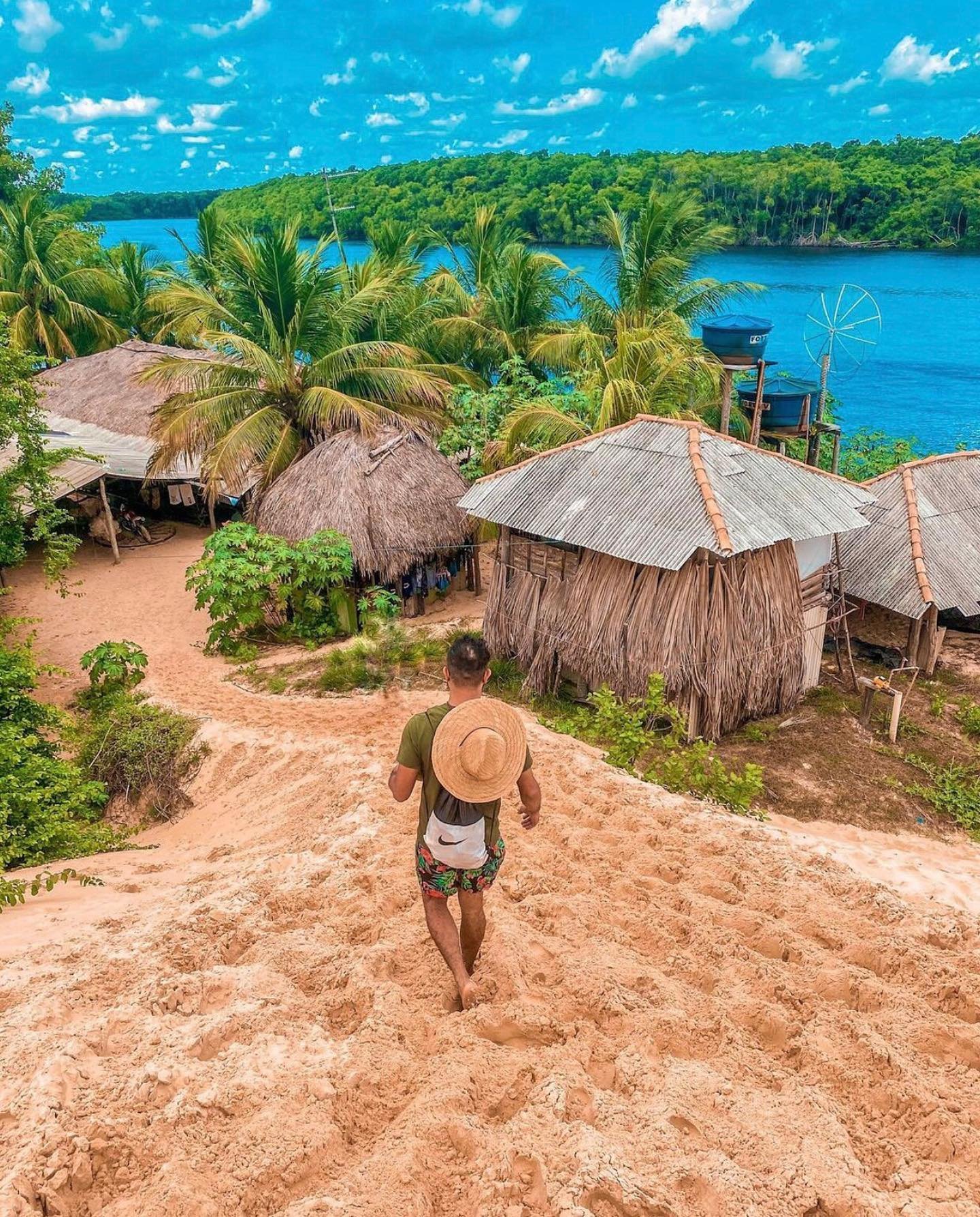 Barefoot into a riverside village — palm shadows, warm sand, and life at the water's edge. A traveller with a straw hat walks barefoot down a sandy slope toward thatched and tin-roofed huts surrounded by palm trees and a vivid blue river on this Lençóis Maranhenses full itinerary