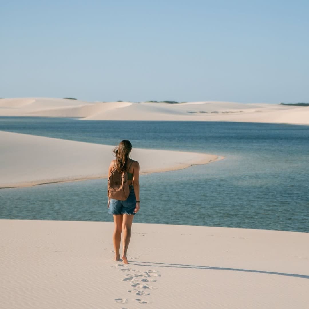 Footprints in the white sand, a lagoon shimmering ahead — this is what the full itinerary feels like. A woman with a backpack walks barefoot across white sand dunes toward a clear turquoise lagoon in Lençóis Maranhenses, her footprints trailing behind her