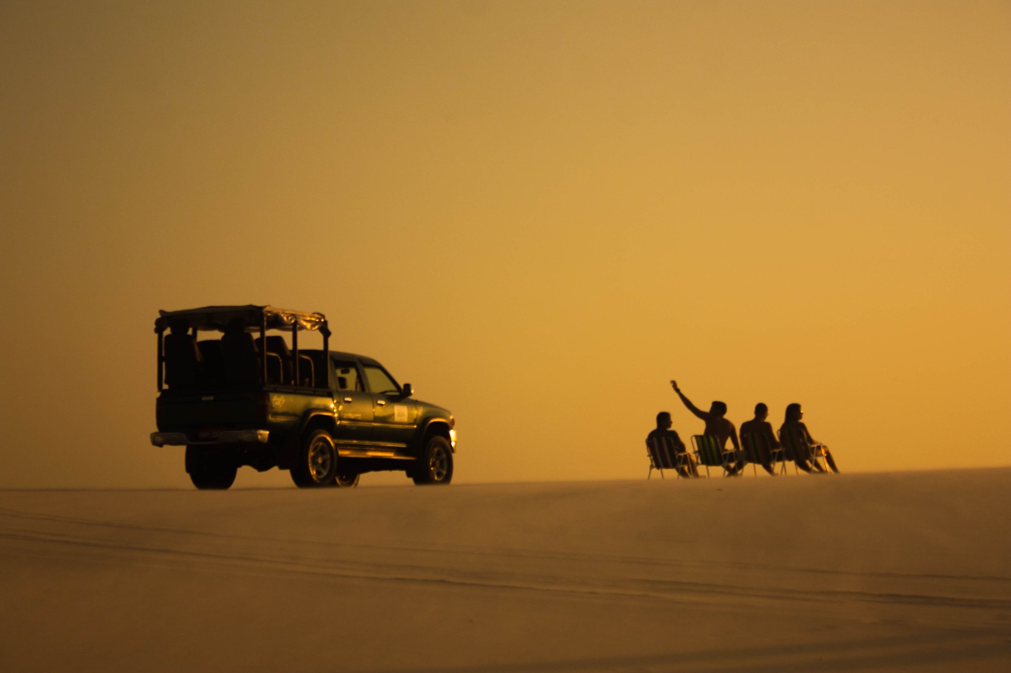 Chairs out, engine off — the dunes belong to everyone at dusk. Photo: Diego Chaves Silhouettes of a 4x4 vehicle and four travellers seated in chairs on vast open sand flats, bathed in deep amber light during a Lençóis Maranhenses full itinerary sunset stop