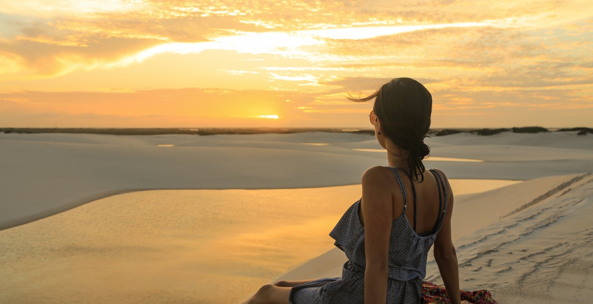 A woman sits on a white sand dune watching a golden sunset over the Lençóis Maranhenses full itinerary landscape of dunes and shallow lagoons