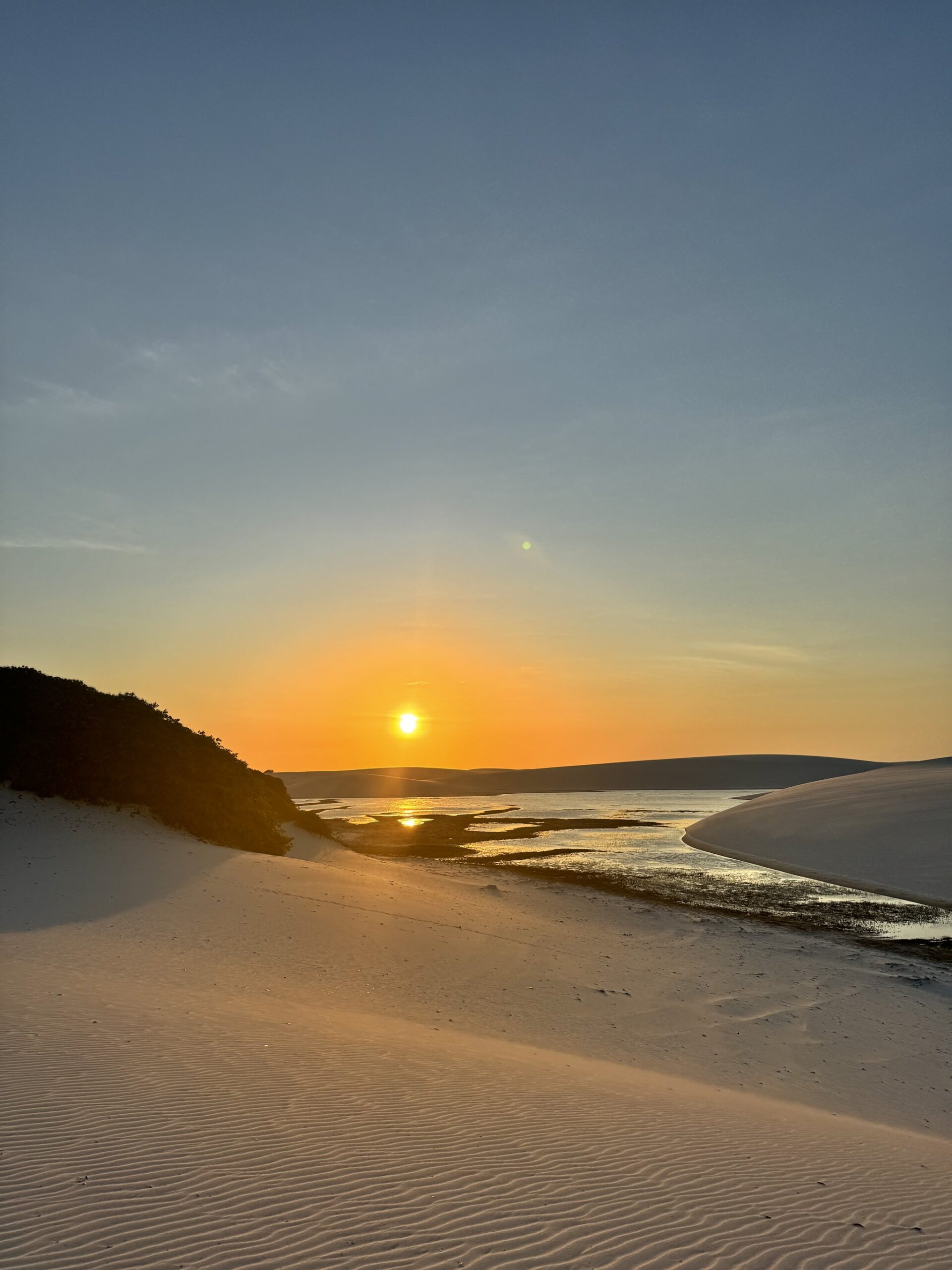 Sun meets water — a molten horizon over white dunes and a still, shallow lagoon. Golden sun setting over a shallow lagoon nestled between white sand dunes on the Lençóis Maranhenses full itinerary route, with rippled sand in the foreground and silhouetted vegetation to the left