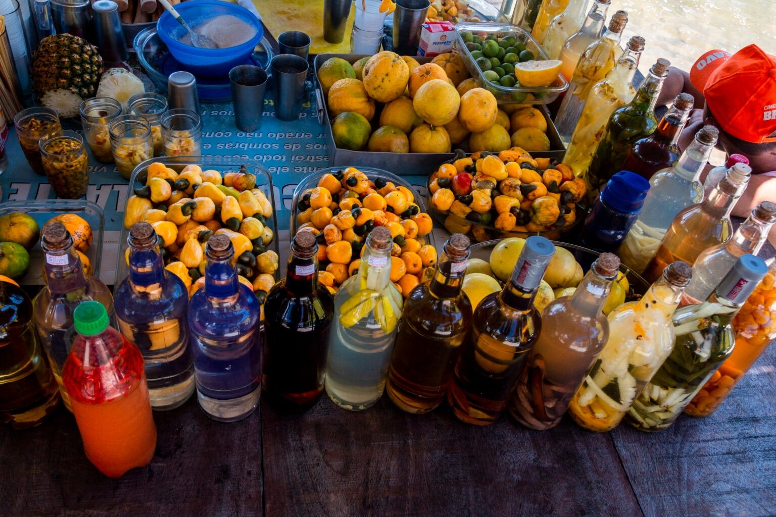 Bottles of every hue — fruit liqueurs and fresh cashew apples at a riverside stop. A market stall spread with trays of fresh cashew fruit and citrus alongside rows of colorful homemade infused liqueur bottles, part of the local food culture on a Lençóis Maranhenses full itinerary