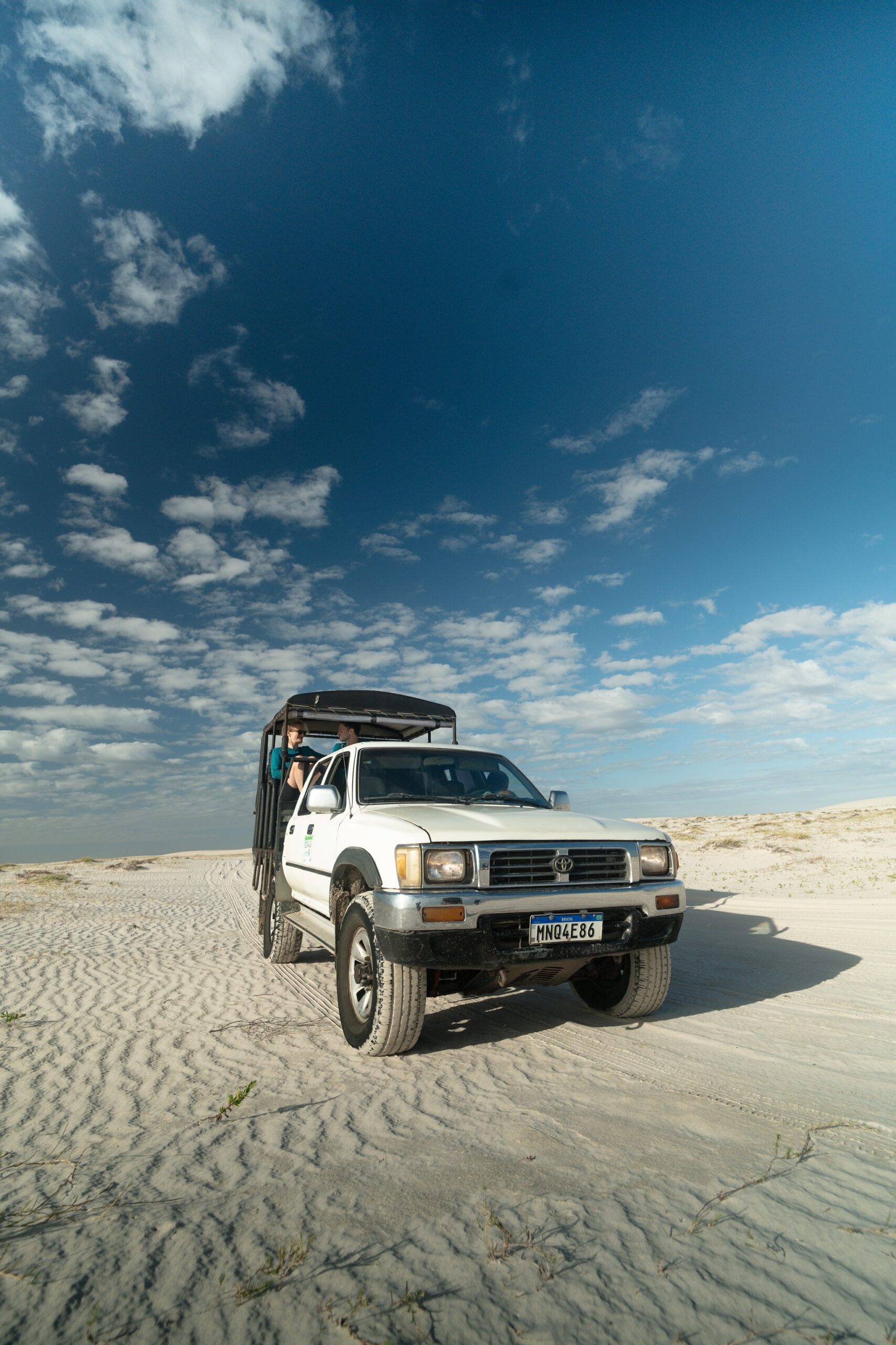 Wind-rippled sand, an open canopy, and the dunes stretching endlessly ahead. A white 4x4 pickup truck with an open canopy carries passengers across the vast white sand dunes of Lençóis Maranhenses under a dramatic blue sky with scattered clouds, part of the Lençóis Maranhenses hike logistics