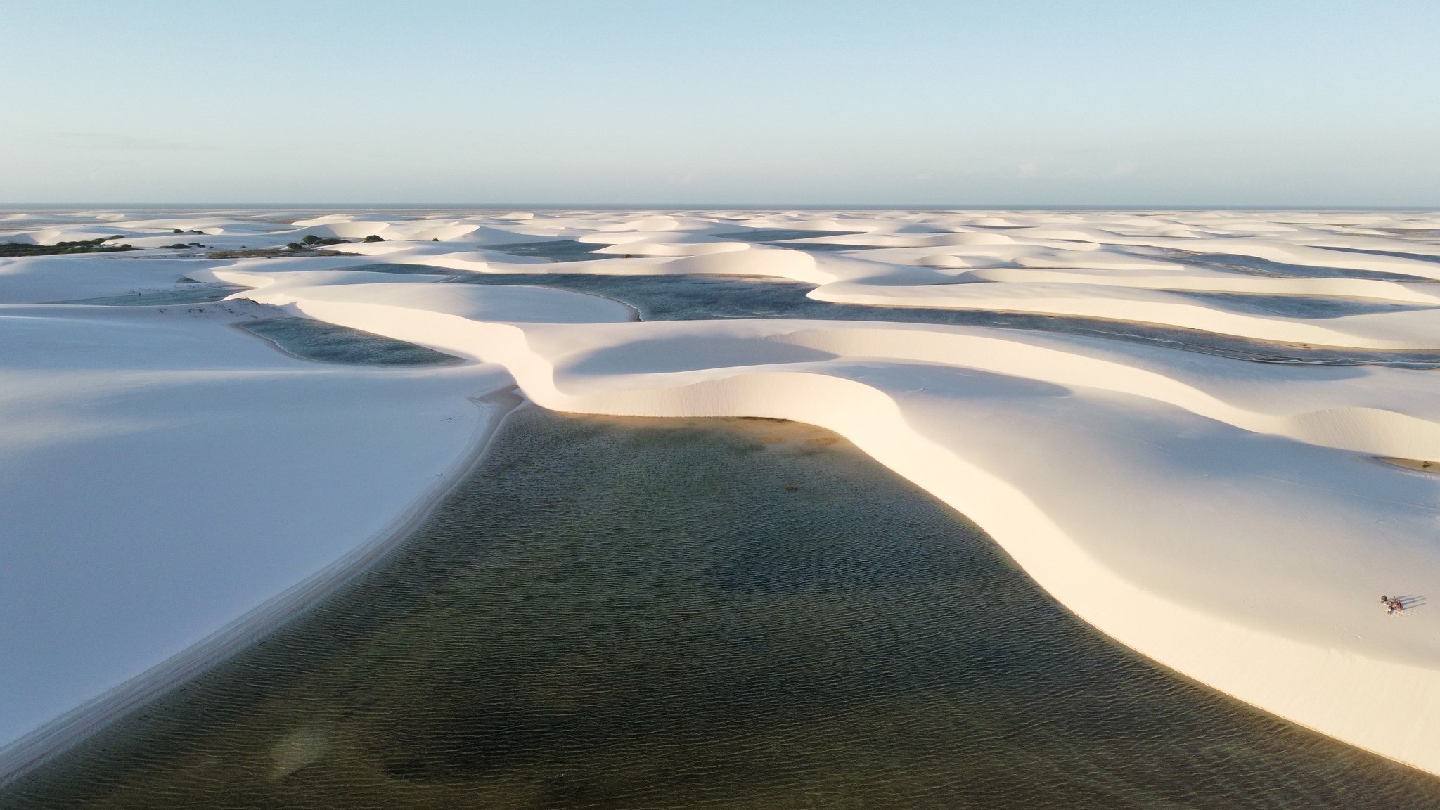 An ocean of white dunes, one green lagoon — the scale of it stuns every time. Aerial view of the Lençóis Maranhenses hike landscape, with vast white sand dunes curving around a dark-green freshwater lagoon stretching to the horizon under a pale blue sky