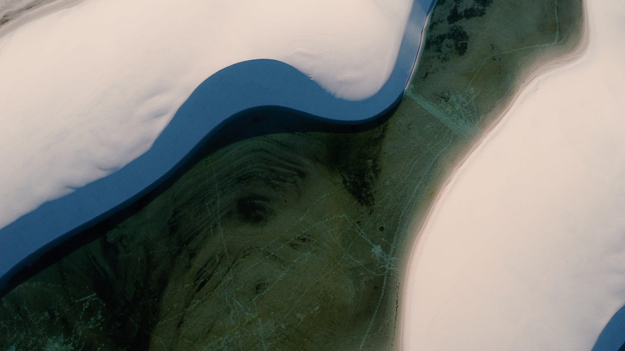 Sand, water, and depth — an abstract canvas seen only from above. Aerial view of a deep-blue lagoon cutting through white sand dunes with submerged green vegetation visible on the Lençóis Maranhenses hike route