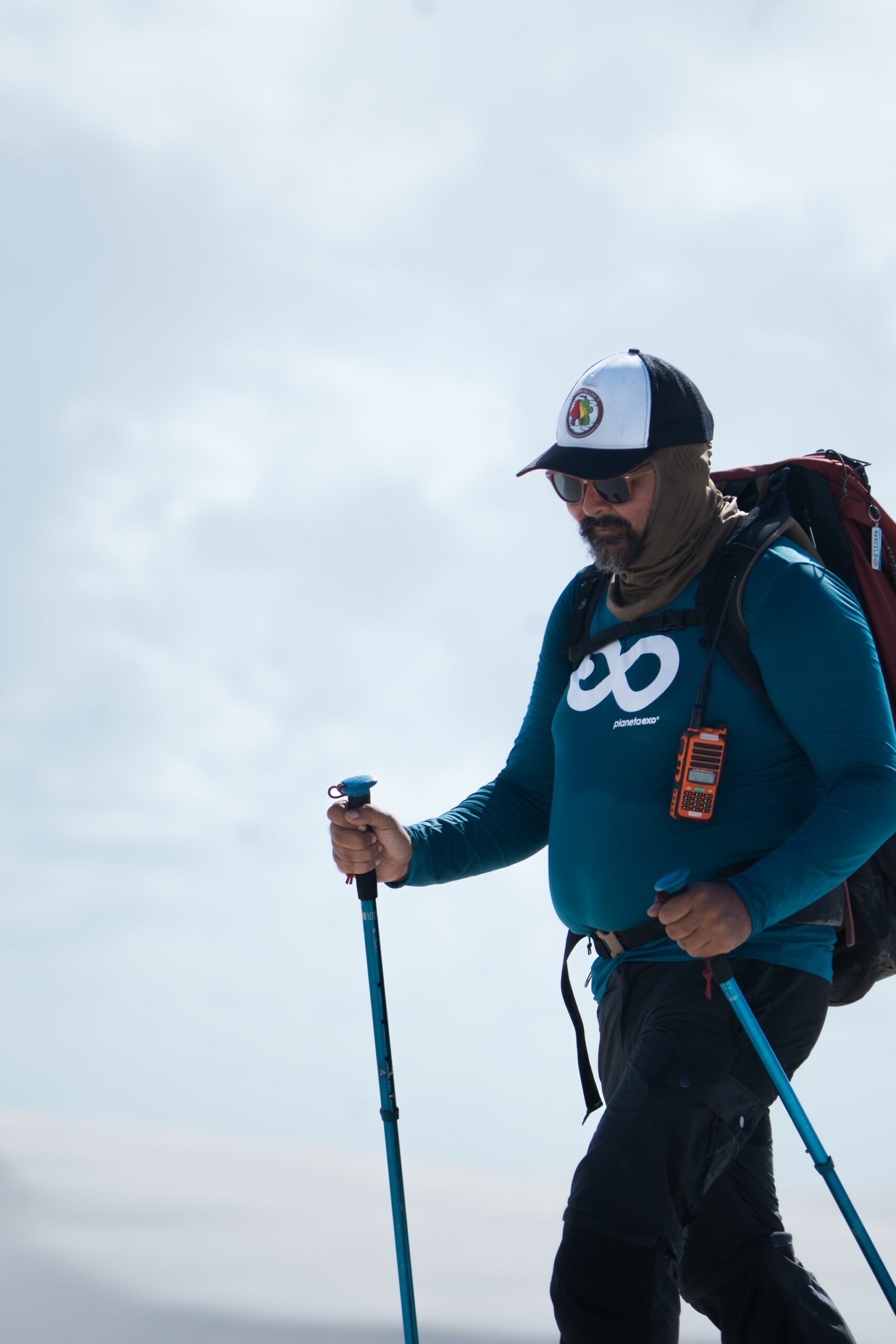 Poles planted, radio clipped, pack loaded — the guide sets the pace. A PlanetaEXO guide equipped with trekking poles, a large backpack, and a radio leads the Lençóis Maranhenses hike under an overcast sky