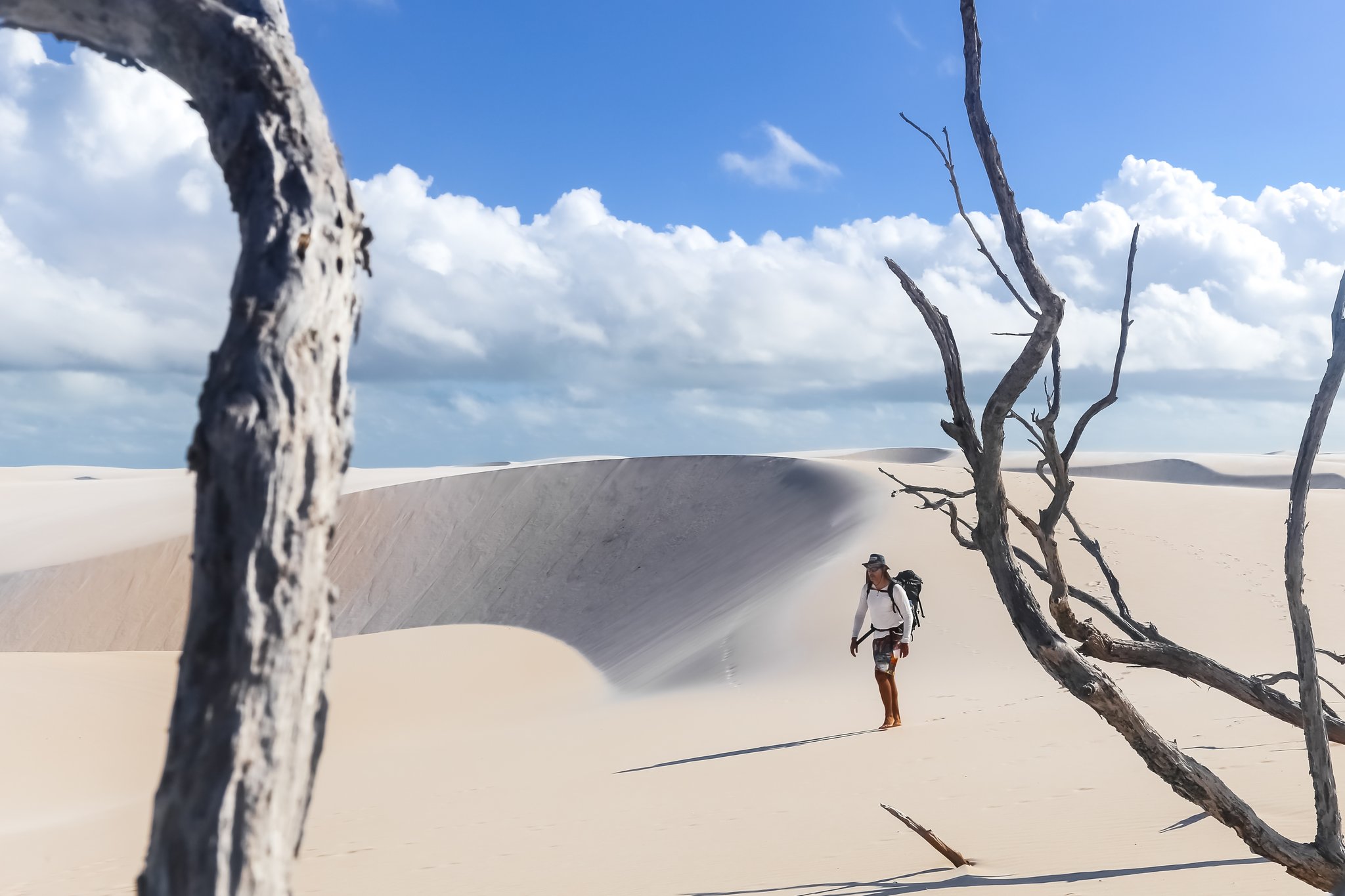Dead wood, white sand, and a lone trekker swallowed by the dunes. Photo: MaRabelo A backpacker wearing a hat pauses among bleached dead trees on a vast white sand dune during a Lençóis Maranhenses hike, with sculpted dune ridges rising under a bright blue sky
