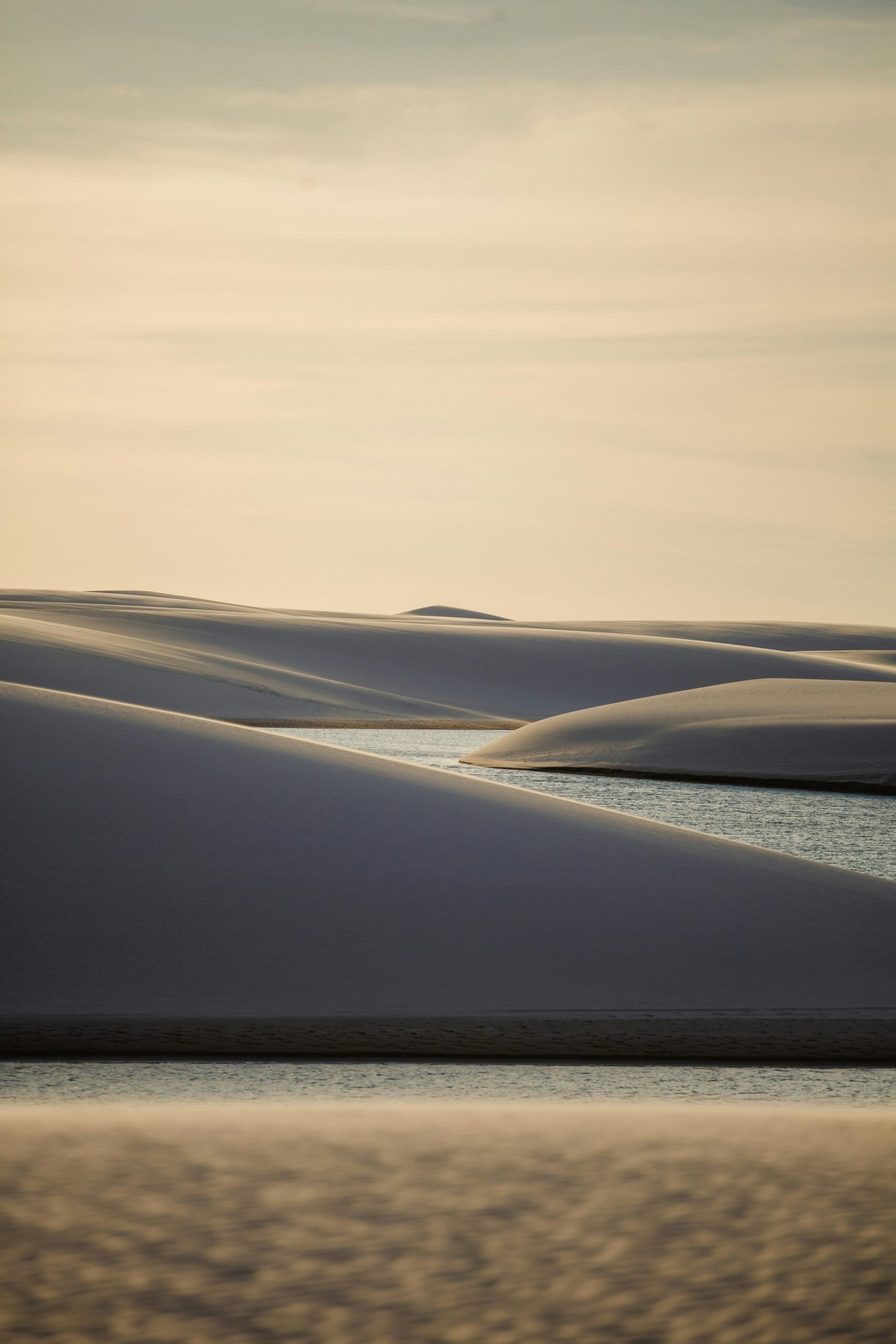 Silence in layers — white dunes, still water, and a pale sky fading to gold. Smooth white sand dunes casting deep blue shadows over still lagoon waters in the soft light of dusk during a Lençóis Maranhenses hike