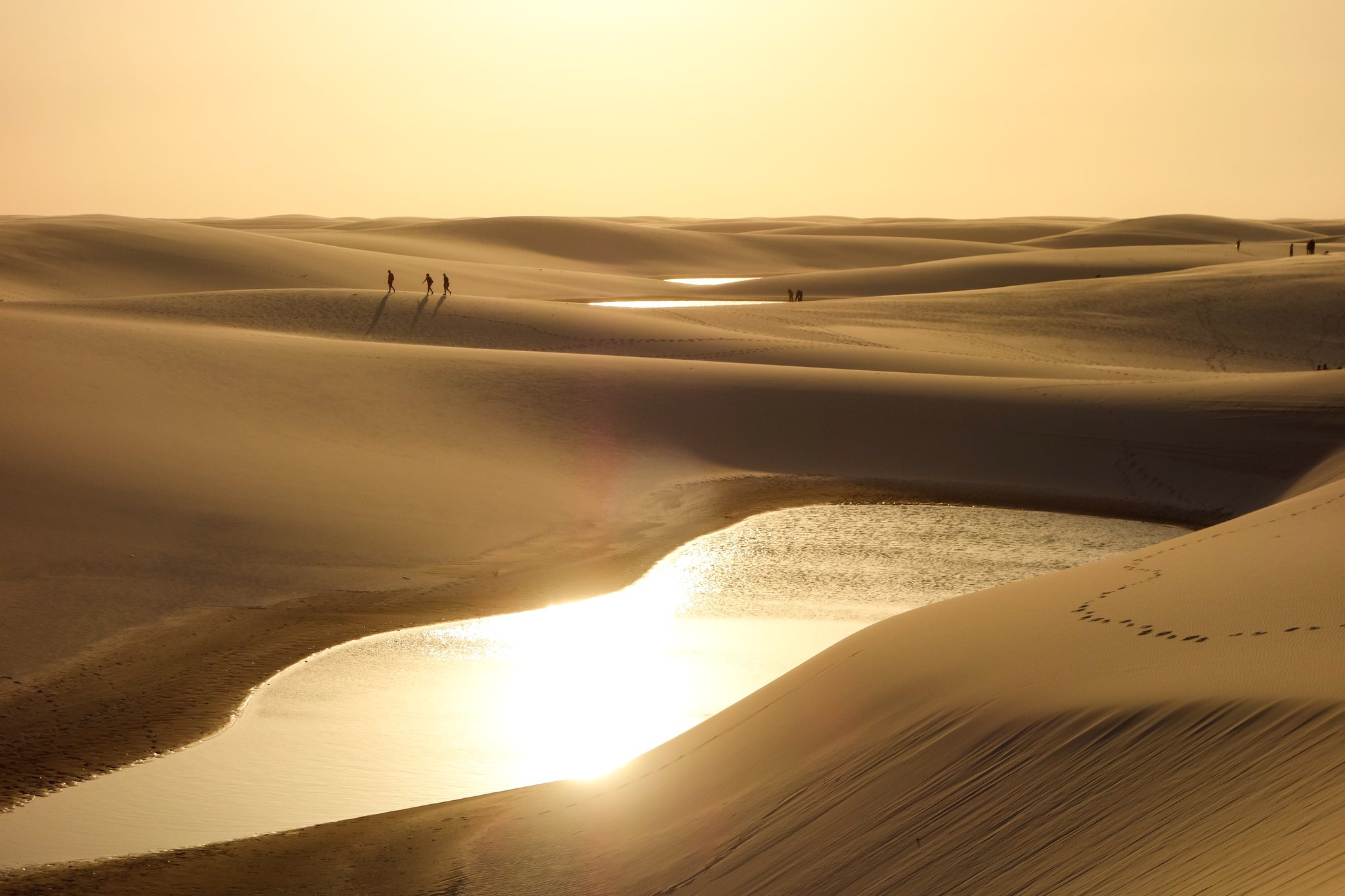 Golden light floods the dunes, and the lagoons burn like mirrors. Photo: caio acquesta Silhouettes of hikers walking across sweeping sand dunes at golden hour during a Lençóis Maranhenses hike, with sunlit freshwater lagoons glowing between the dunes in the foreground