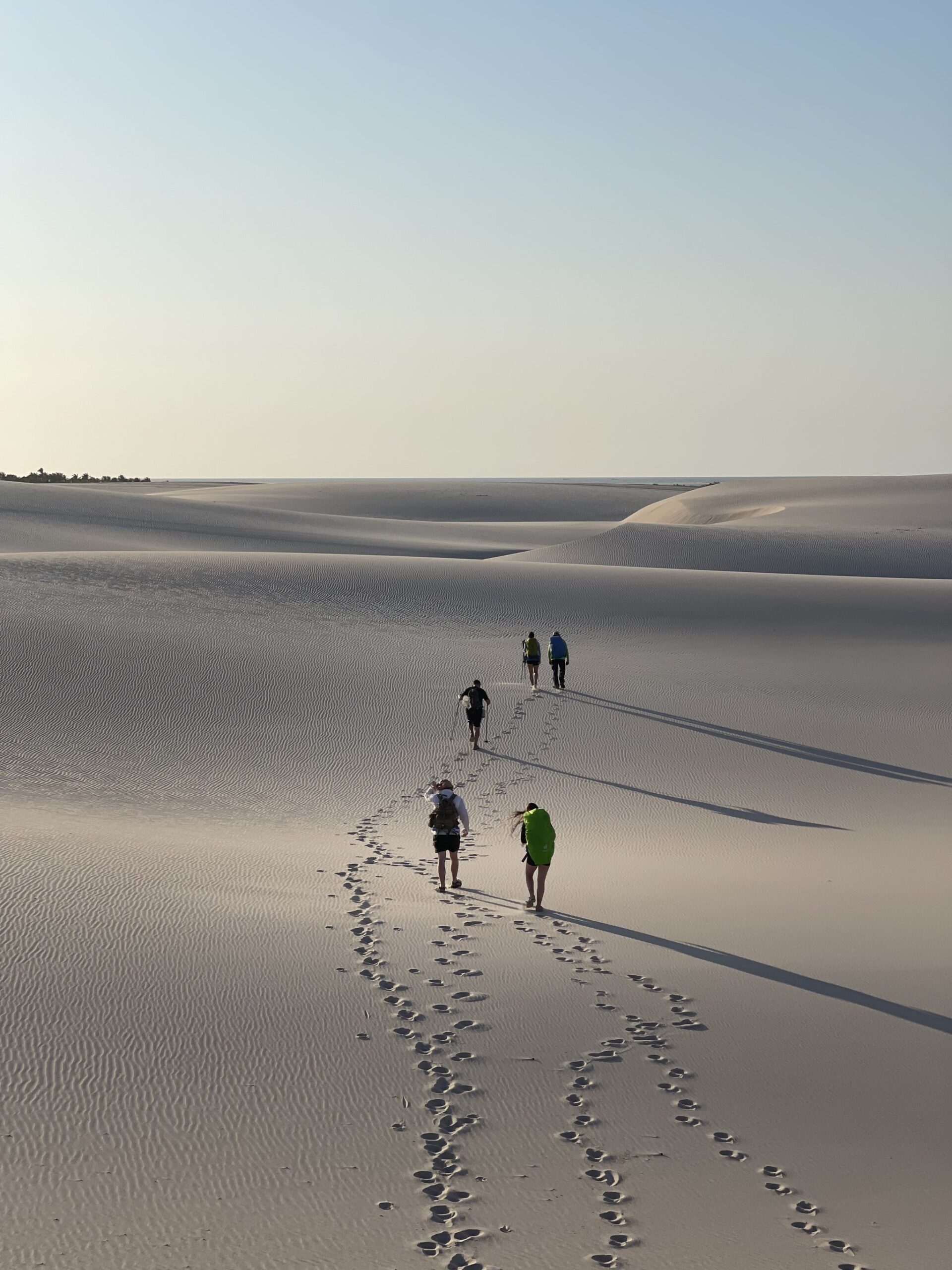 Four packs, one trail pressed into the sand, and nothing but dunes to the horizon. Four hikers with backpacks trace a line of footprints across vast rippled white sand dunes on the Lençóis Maranhenses hike, under a pale morning sky