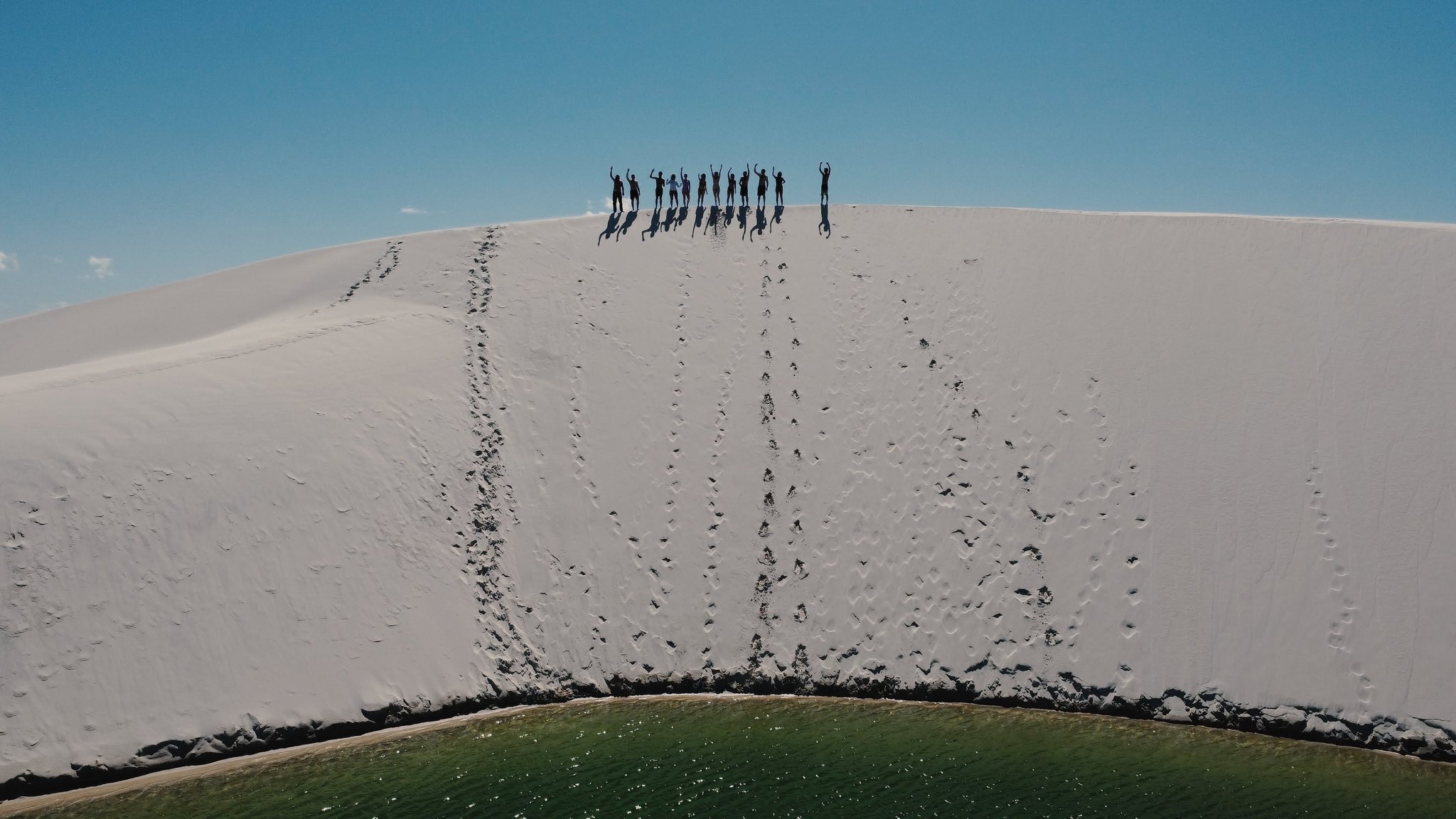 Twelve silhouettes, arms raised, owning the top of the dunes. A group of hikers silhouetted against a blue sky atop a vast white sand dune during a Lençóis Maranhenses hike, with a green lagoon visible at the dune's base and footsteps trailing down the face