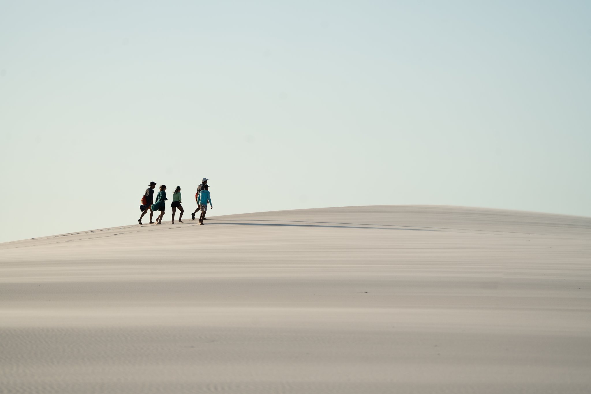 Four silhouettes, one endless dune — the scale of the Lençóis speaks for itself. Four hikers walking along the crest of a vast white sand dune during a Lençóis Maranhenses hike, tiny figures against a pale open sky