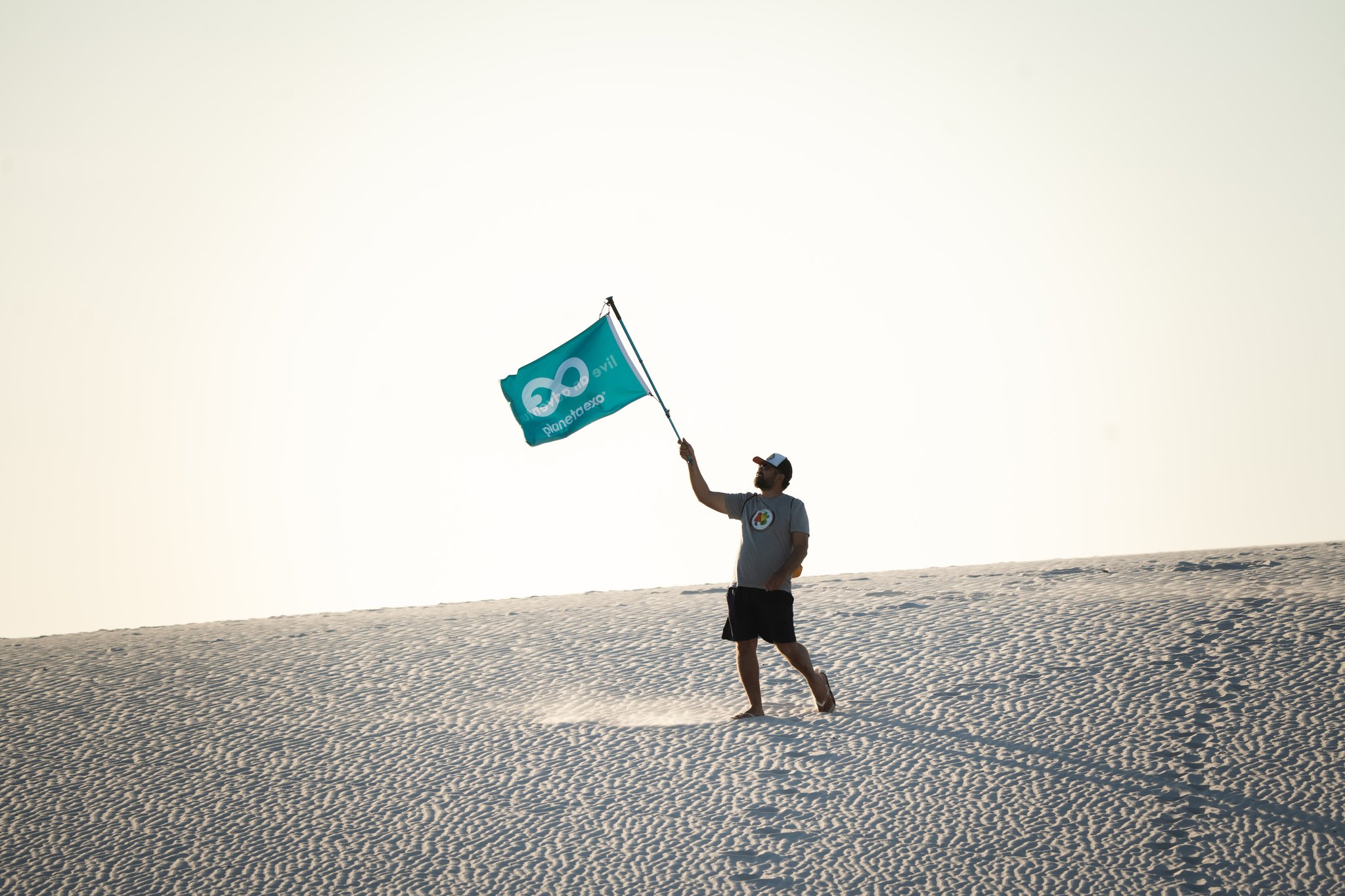 A guide waves a teal PlanetaEXO flag while striding across vast white sand dunes on a Lençóis Maranhenses hike