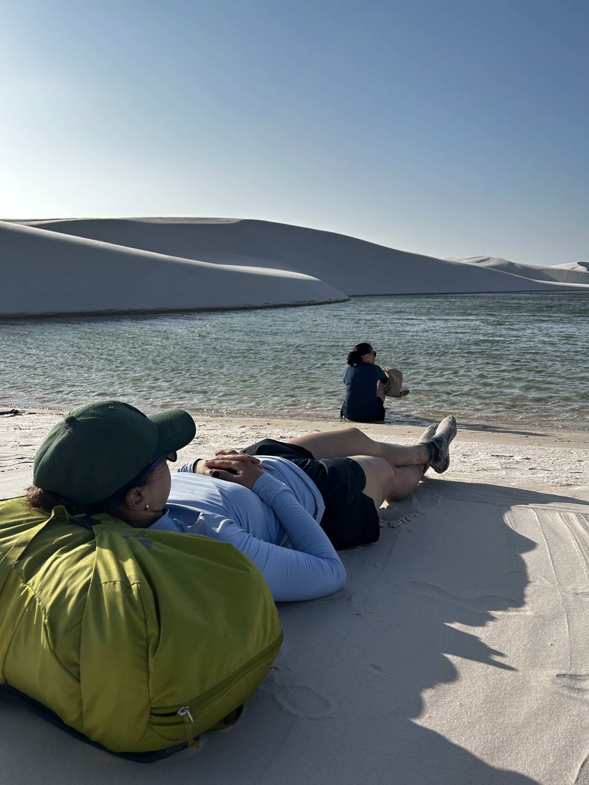Bare feet, white dunes, a lagoon — rest earned. A hiker rests on white sand dunes beside a clear freshwater lagoon during a Lençóis Maranhenses hike, while another traveller sits at the water's edge with rolling white dunes beyond