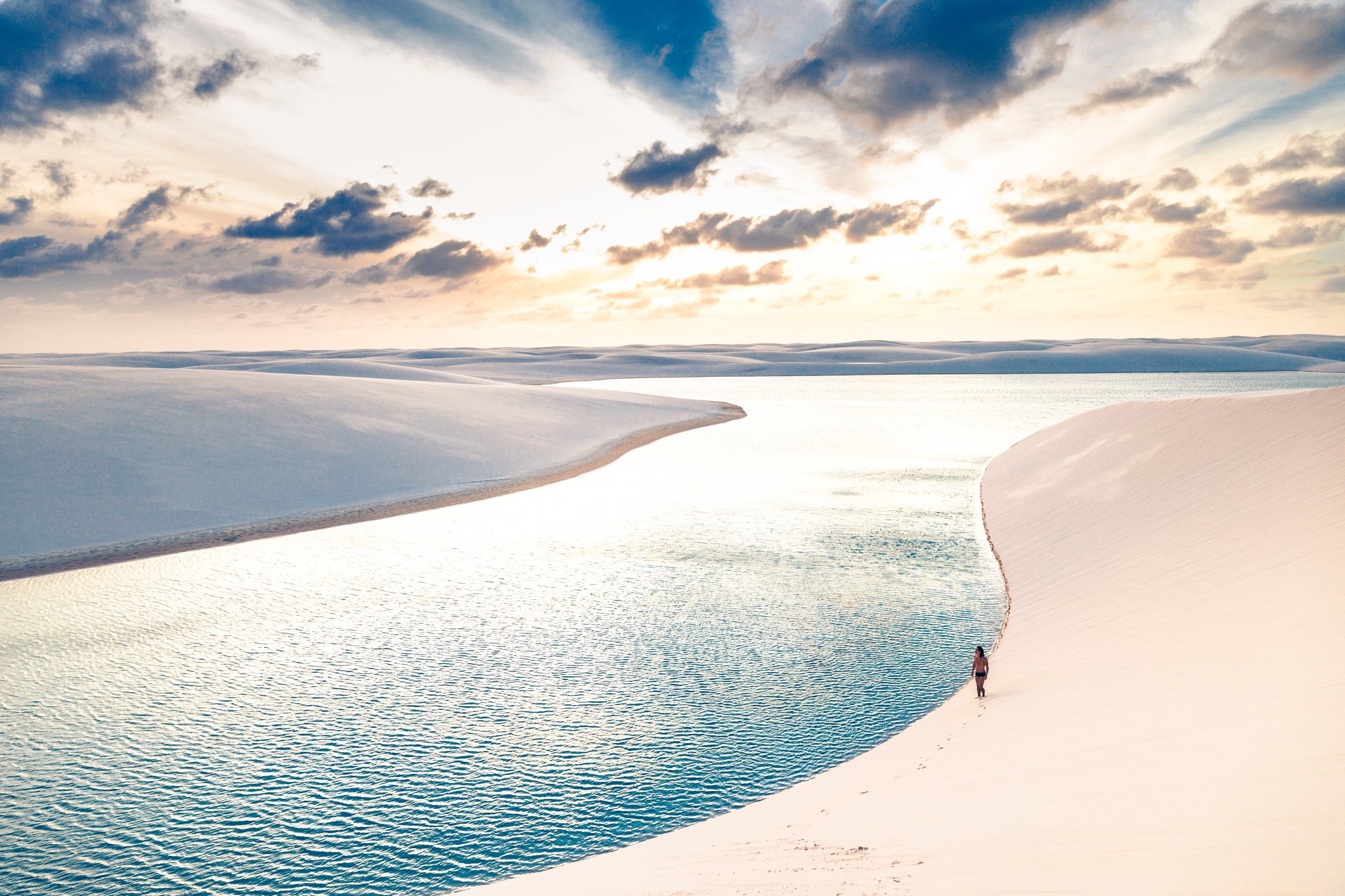 One person, one lagoon, endless white dunes stretching to the horizon. Photo: cokada A lone walker traces the curved edge of a crystal-clear freshwater lagoon between sweeping white dunes during a Lençóis Maranhenses hike, under a dramatic sunset sky