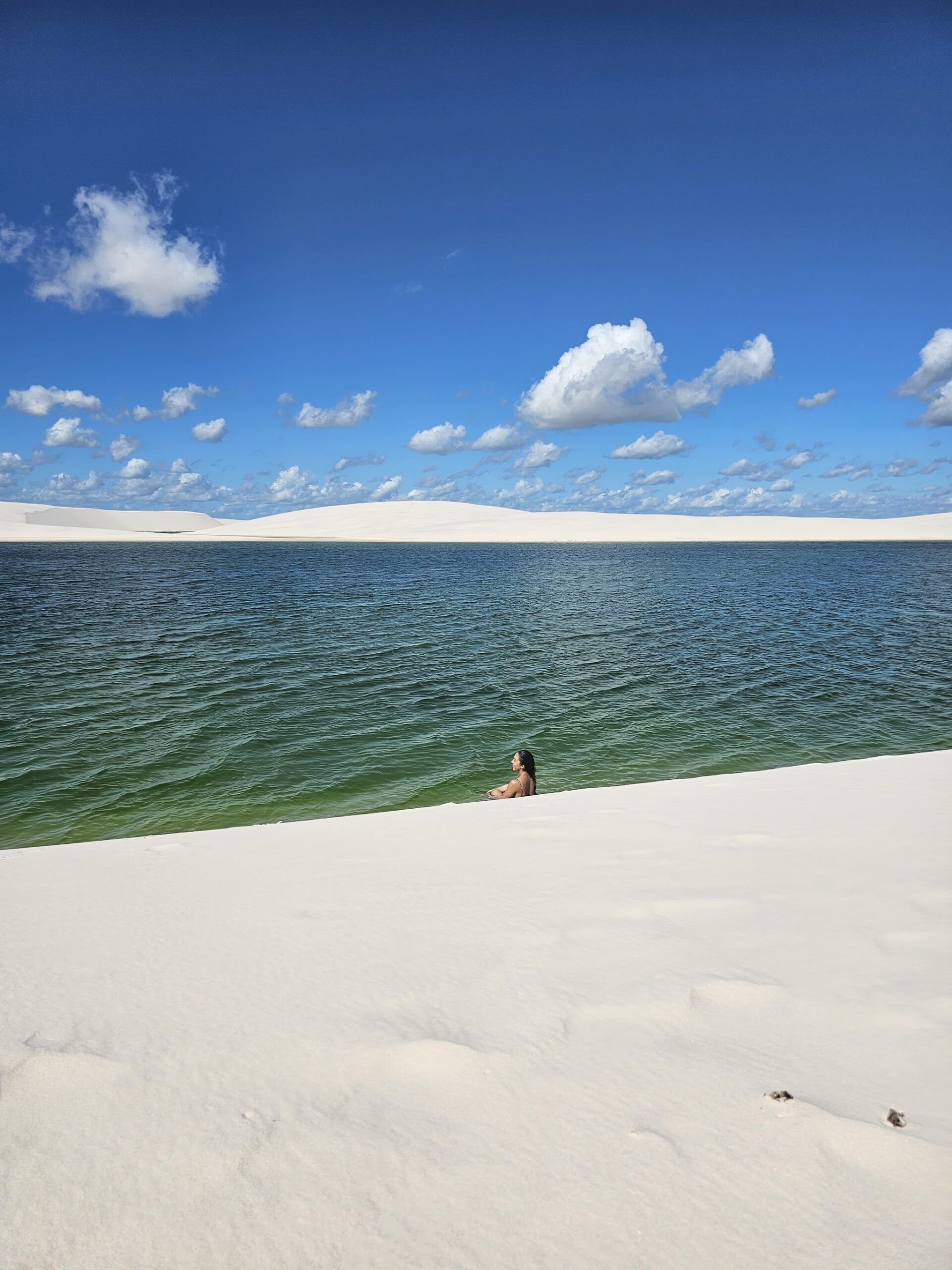 Adrift in green water, white dunes rising all around. A lone swimmer waist-deep in a wide green freshwater lagoon on the Lençóis Maranhenses hike, framed by smooth white dunes and a vivid blue sky