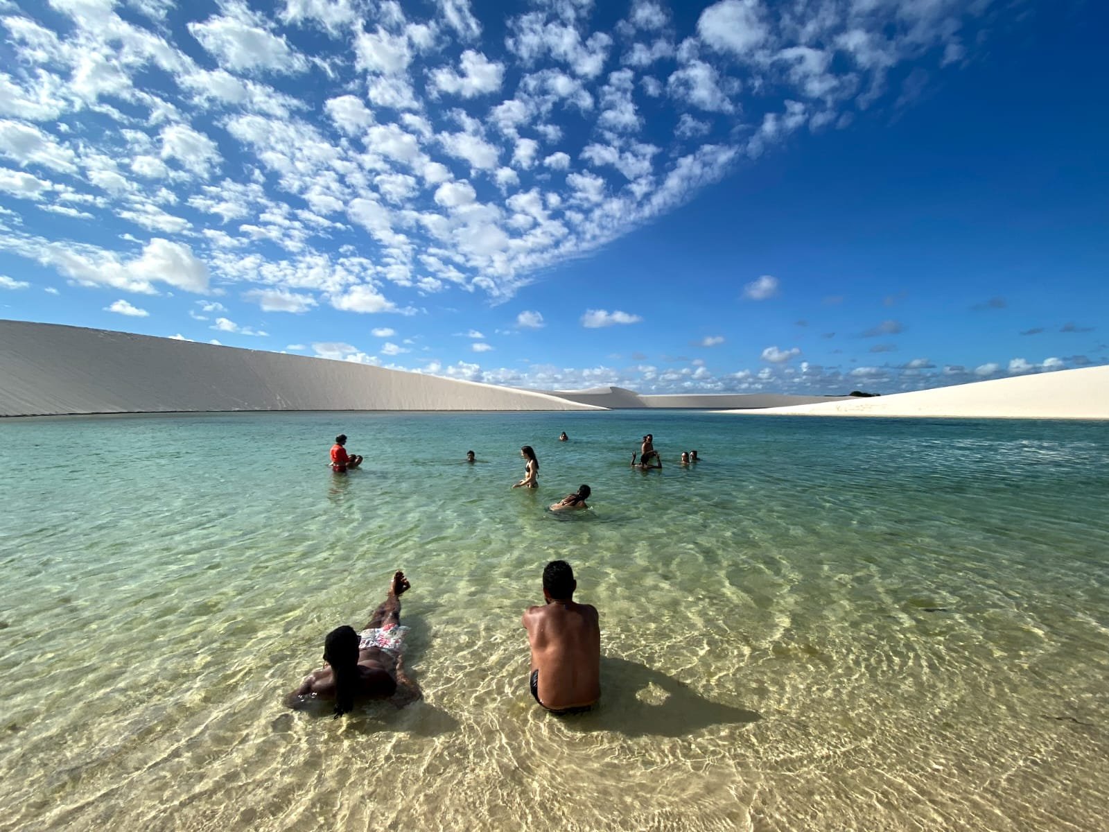 Cool water, warm sand, and absolutely nowhere else to be. A group of travellers wading and relaxing in a crystal-clear freshwater lagoon during a Lençóis Maranhenses hike, surrounded by towering white sand dunes under a vivid blue sky