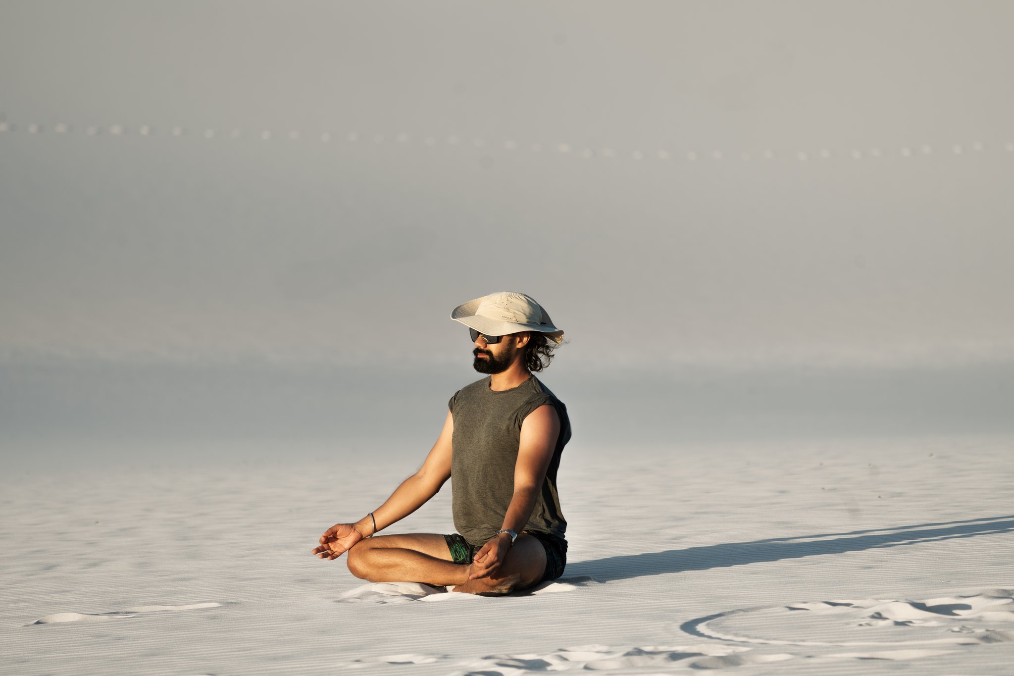 Still and small against the infinite white — a moment of pure stillness mid-trek. A bearded man in a cap and sunglasses sits cross-legged in a lotus pose on the vast white sand flats during a Lençóis Maranhenses hike, surrounded by an endless pale horizon