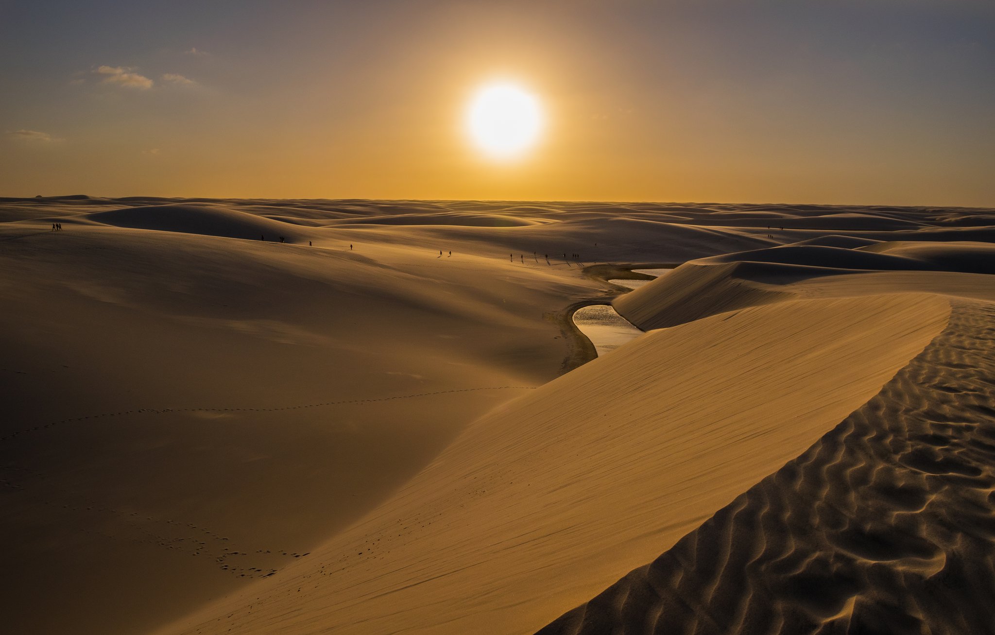 Sun low, dunes molten gold, a ribbon of water curling between ridges. Photo: NidoHuebl Vast rolling white sand dunes of Lençóis Maranhenses stretch to the horizon at golden sunset, with a small serpentine lagoon nestled between the dunes and tiny silhouettes of hikers visible in the distance — a defining scene of the Lençóis Maranhenses hike