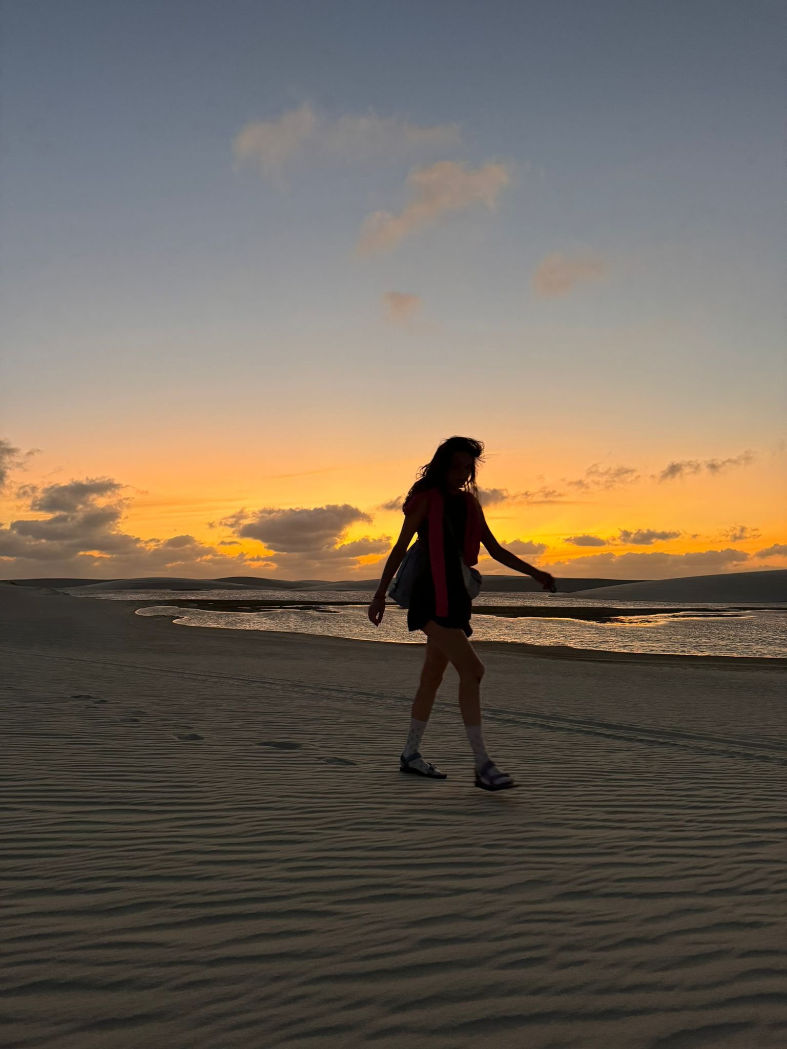 Orange sky, rippled sand — the dunes at their most cinematic. A woman's silhouette walks across wind-rippled sand dunes during a Lençóis Maranhenses hike, a glowing orange sunset and calm lagoon stretching behind her