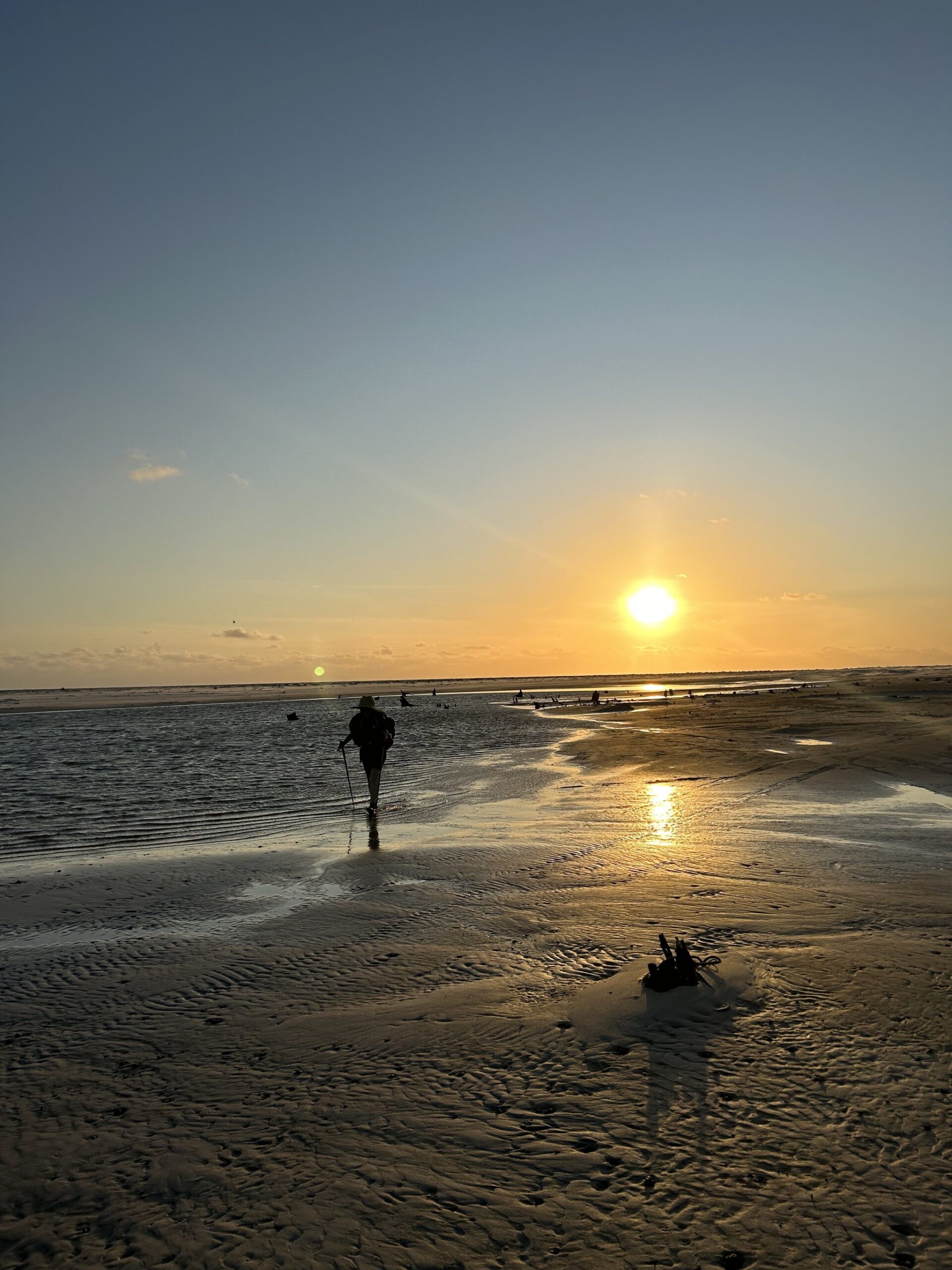 Golden hour, wet sand underfoot, and nowhere left to be but here. A lone hiker with a backpack and trekking poles wades through shallow tidal water on the Lençóis Maranhenses hike, silhouetted against a golden sunset over vast rippled sand flats