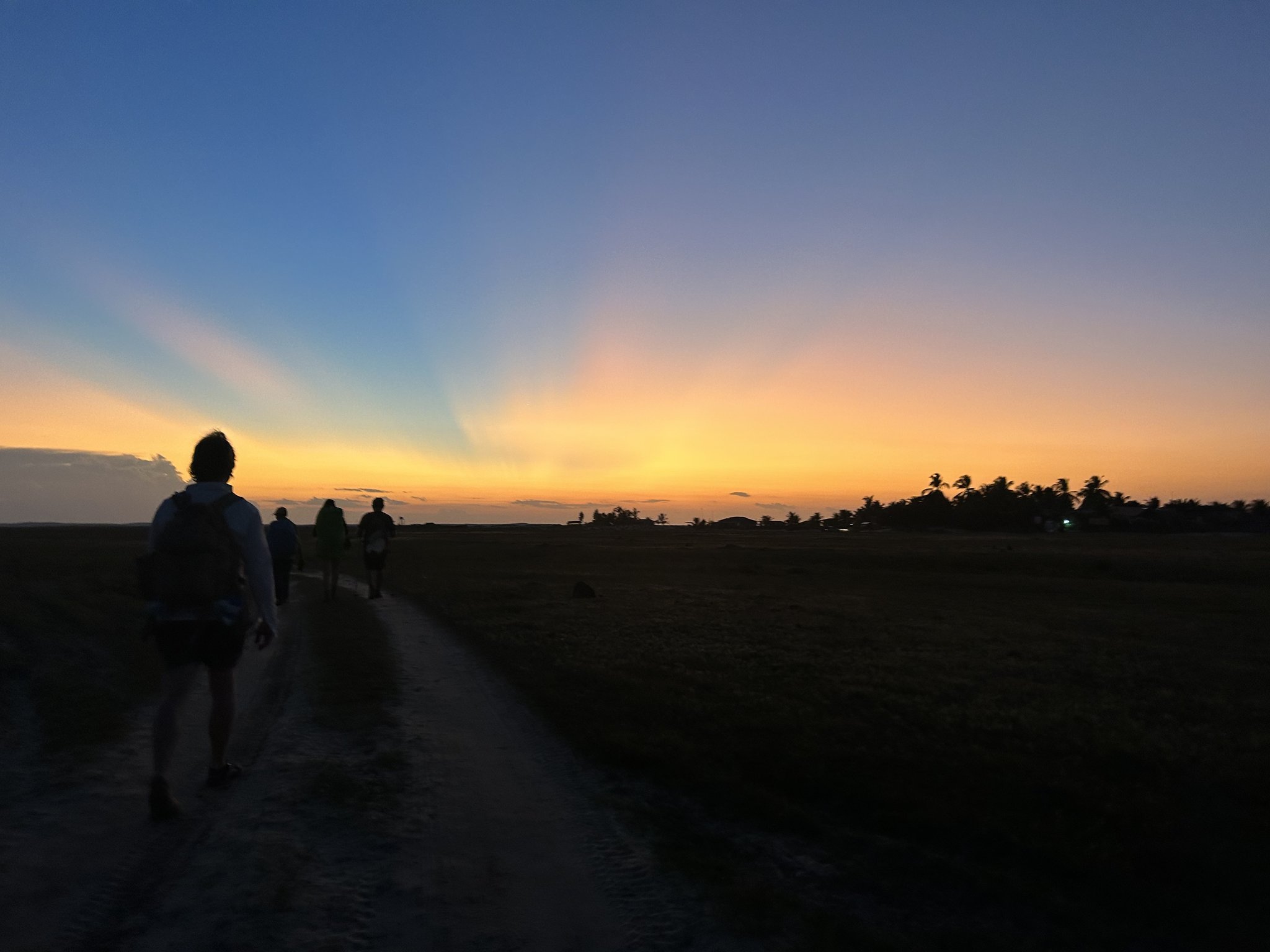 Dusk swallows the plain, and the trail stretches on. Silhouettes of hikers walking a sandy trail toward a vivid orange and blue sunset on the Lençóis Maranhenses hike, with palm trees on the distant horizon