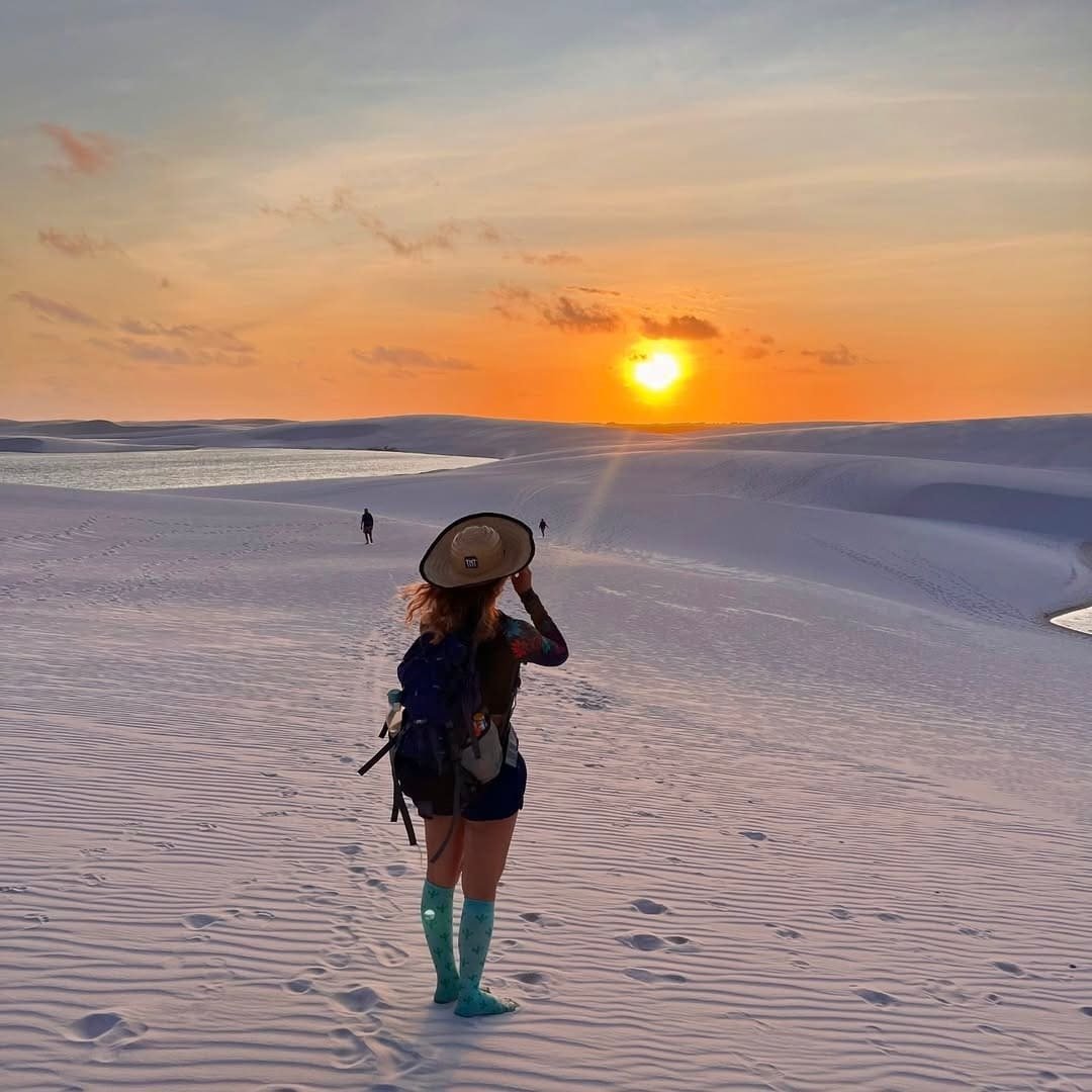 Hat clutched against the wind, the whole horizon on fire. A solo hiker with a backpack and wide-brim hat stands on vast white dunes during the Lençóis Maranhenses hike, facing a blazing orange sunset with a freshwater lagoon glimmering in the distance