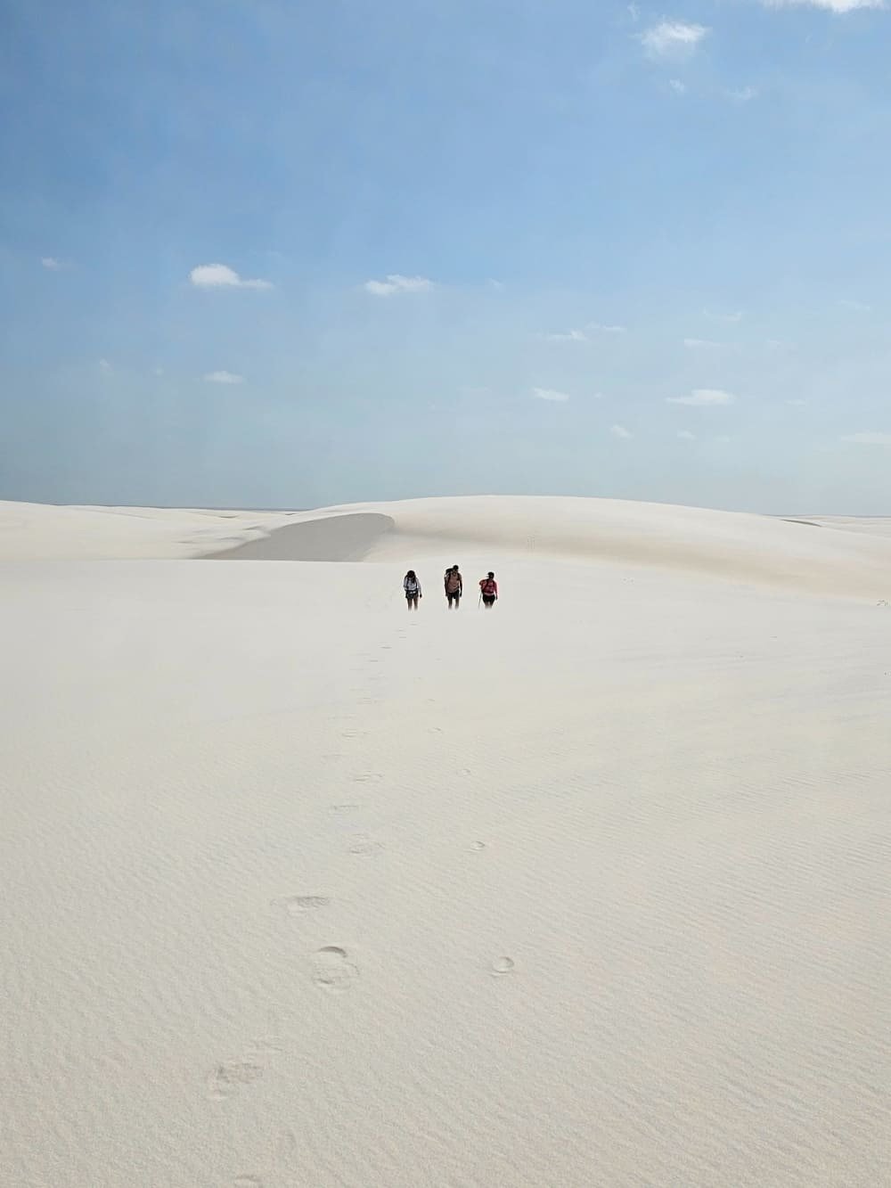 Footprints in the white — three trekkers disappearing into the dunes. Three hikers walking across vast white sand dunes on a Lençóis Maranhenses hike, their footprints trailing behind them toward a pale blue sky