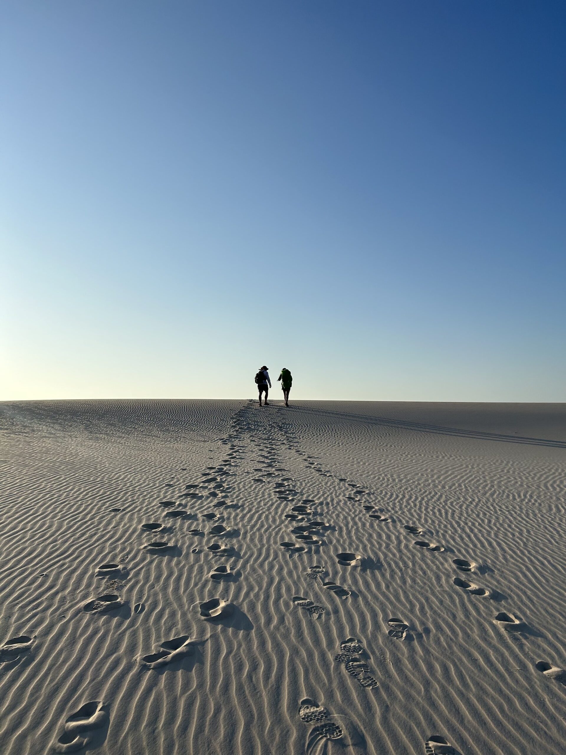 Just footprints, endless sand, and the horizon pulling you forward. Two silhouetted hikers with backpacks walk toward the horizon along a vast rippled white sand dune on a Lençóis Maranhenses hike, their footprints trailing back across the foreground under a clear blue sky