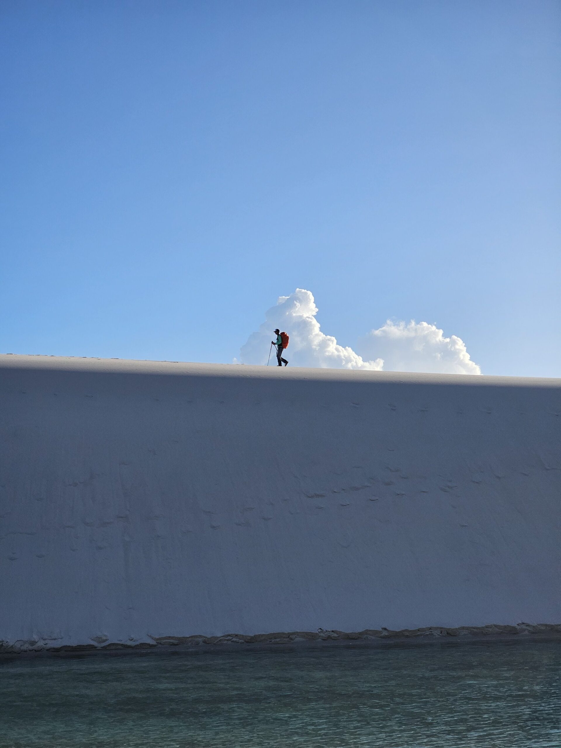 Small against the white — a single ridge between two worlds. A lone hiker with a red pack and trekking pole silhouetted on the razor-sharp crest of a brilliant white sand dune during the Lençóis Maranhenses hike, with turquoise lagoon water visible at the base and a towering cumulus cloud rising behind