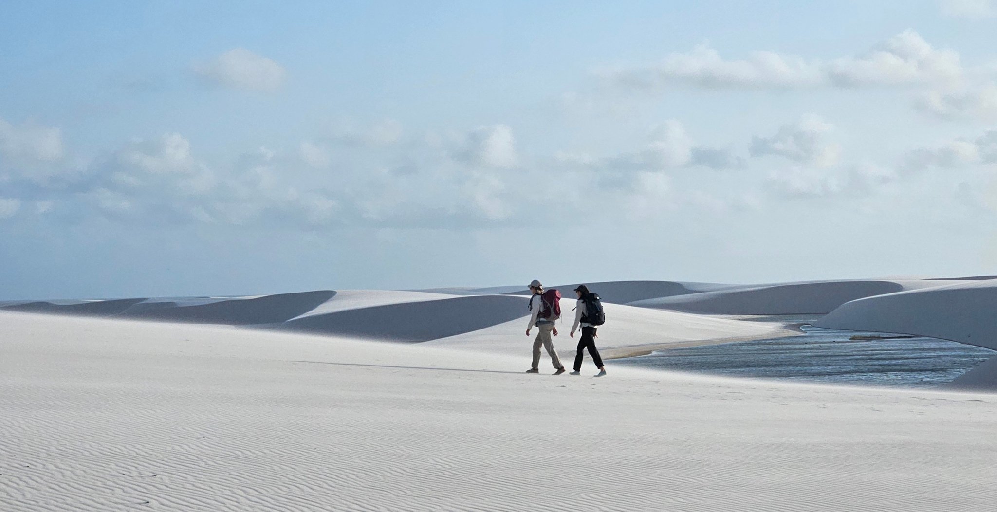 Two hikers with backpacks walk across vast white sand dunes on the Lençóis Maranhenses hike, with a shallow lagoon and rolling dunes stretching to the horizon under a pale blue sky