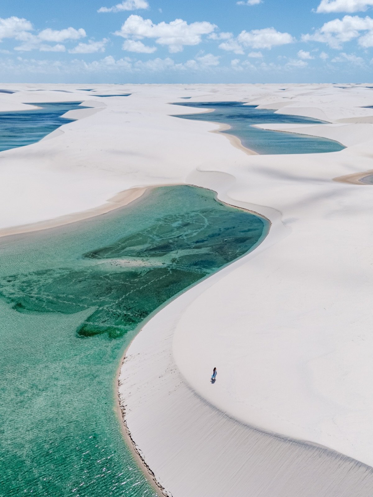 One person, endless white dunes, and water the color of sea glass. Aerial view of Lençóis Maranhenses Santo Amaro showing vast white sand dunes dotted with vivid turquoise and emerald rainwater lagoons stretching to the horizon, with a lone figure standing on a dune ridge beside a large clear lagoon