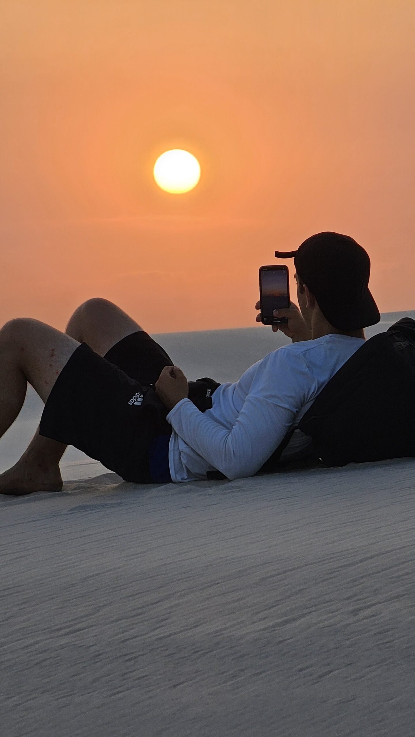 Stretched out on the dunes, phone raised — some sunsets demand a second look. A traveller reclines on white sand dunes in Lençóis Maranhenses Santo Amaro, photographing a vivid orange sunset with a smartphone