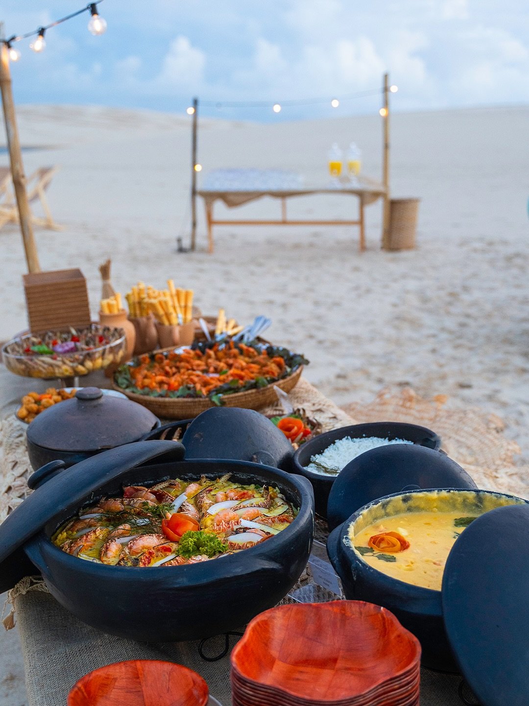 Clay pots, candlelight, and the dunes stretching endlessly behind — dinner tastes different out here. Multiple black clay pots filled with shrimp stew and creamy seafood dishes arranged on a rustic table on white sand dunes at dusk, with string lights glowing in the background near Lençóis Maranhenses Santo Amaro