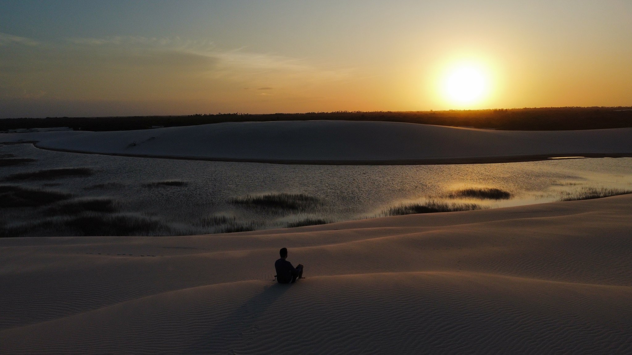 Quiet enough to hear the wind shift — sunset over the lagoons. A lone traveller sits silhouetted on white sand dunes overlooking a still lagoon at sunset in Lençóis Maranhenses Santo Amaro, with golden light reflecting off the water